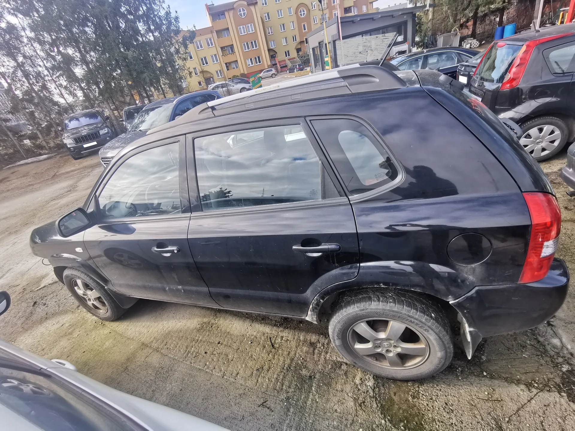 Czarny samochód typu SUV zaparkowany na otwartym placu parkingowym. W tle widoczne są inne pojazdy, budynki mieszkalne, drzewa oraz niebo. Samochód ma czarną karoserię, srebrne felgi i czerwone światła tylne. Plac parkingowy ma nierówną, brudną nawierzchnię. W tle stoją również inne samochody, w tym ciemny SUV i czarny hatchback.