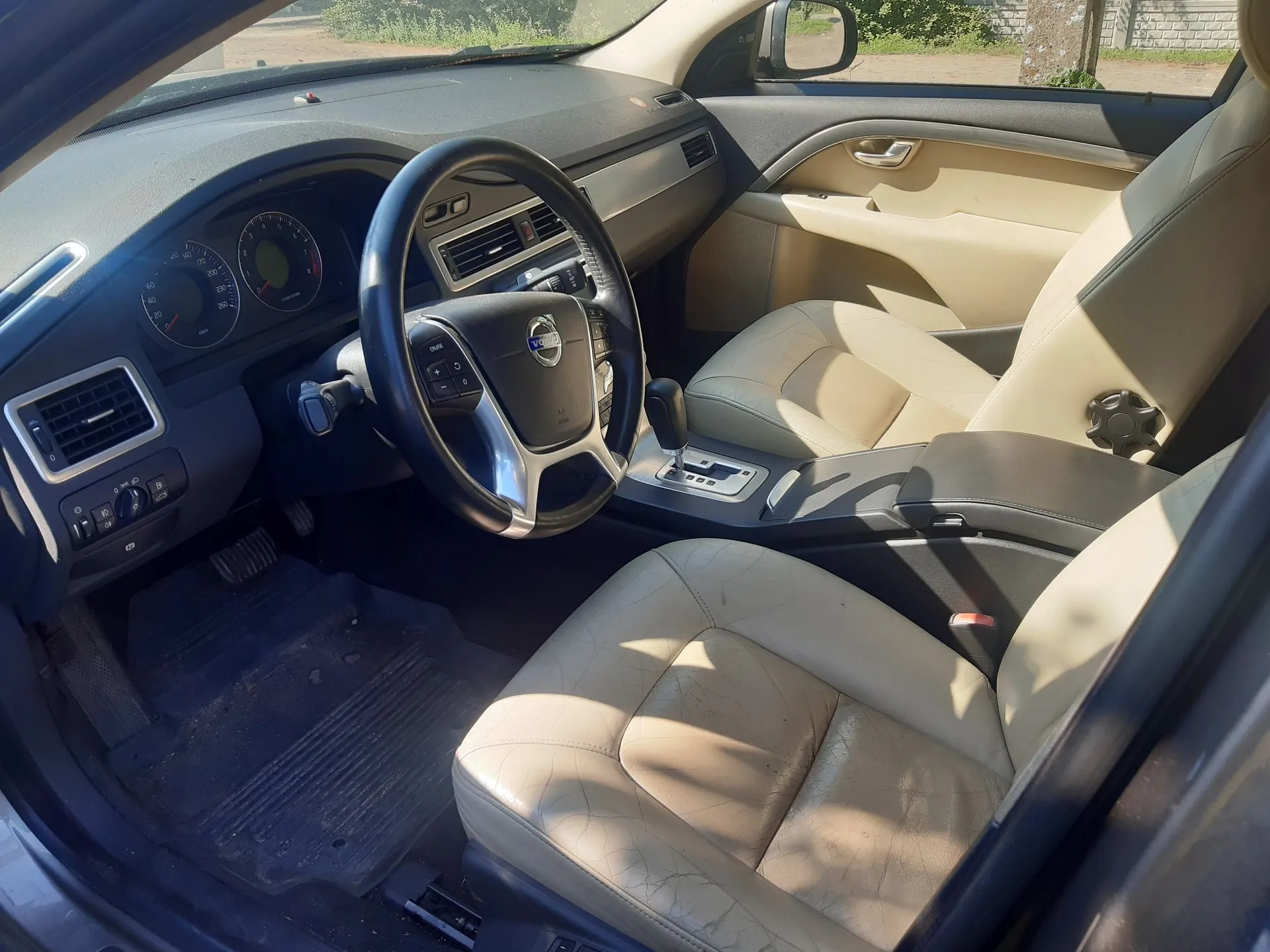 Wnętrze samochodu z widokiem na stronę kierowcy. Widoczna jest czarna kierownica z logo Volvo, deska rozdzielcza z zegarami i nawiewami oraz beżowe, skórzane fotele. Samochód ma ciemne wykończenie wnętrza i jasną tapicerkę siedzeń.