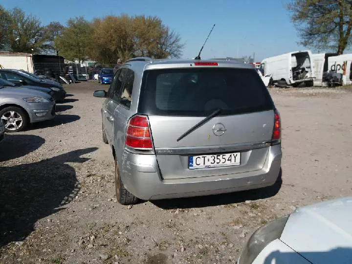 Srebrny samochód osobowy marki Opel, widok od tyłu, z tablicą rejestracyjną CT 3554Y. Po lewej stronie inne zaparkowane pojazdy, w tle biały furgon z napisem DPD oraz drzewa. Teren parkingowy z żwirem, jasne niebo. Samochód stoi na płaskim obszarze, bez widocznych osób.