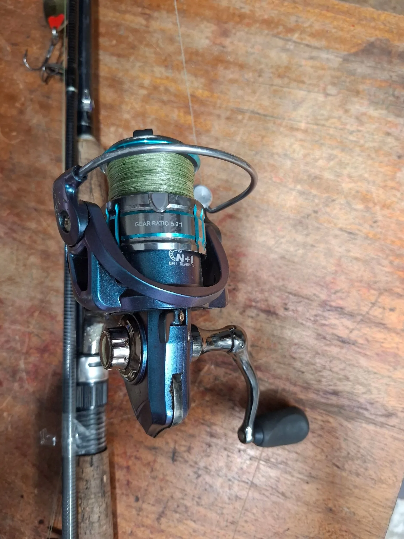 Zbliżenie na niebieski kołowrotek wędkarski zamontowany na wędce, z nawiniętą zieloną żyłką. Na szpuli widnieją napisy: 'GEAR RATIO 5.2:1' oraz 'N+1 BALL BEARINGS'. Kołowrotek posiada metalową korbkę z czarnym uchwytem. Całość spoczywa na drewnianym blacie.