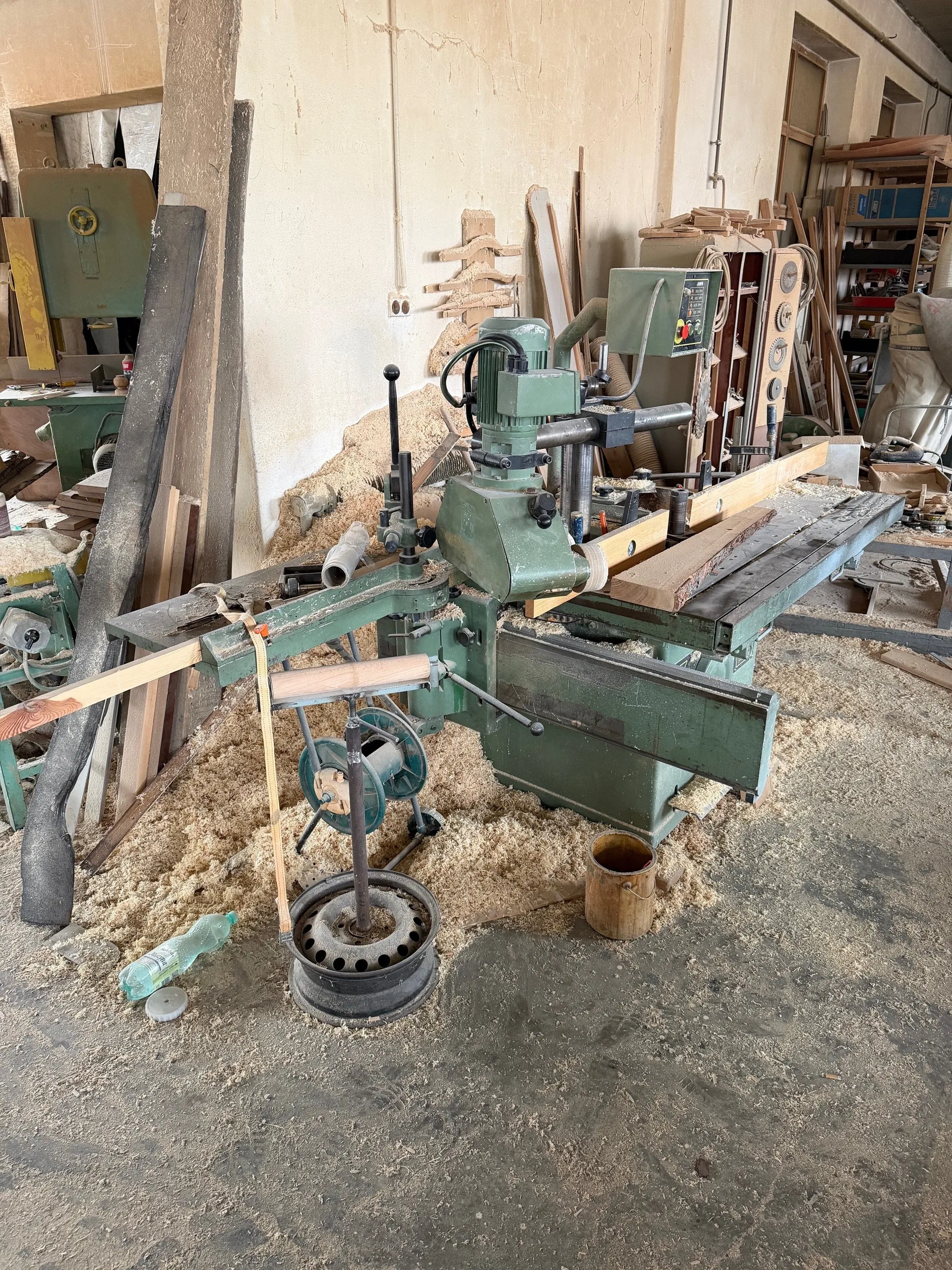 Wnętrze warsztatu stolarskiego z zielonym maszynami do obróbki drewna. Na podłodze i maszynach widoczny jest trocin. Wokół znajdują się kawałki drewna, butelka, wiadro oraz półki z narzędziami i pudełkami. Ściany pomieszczenia są zużyte, a na tle widoczne są dodatkowe urządzenia i materiały.