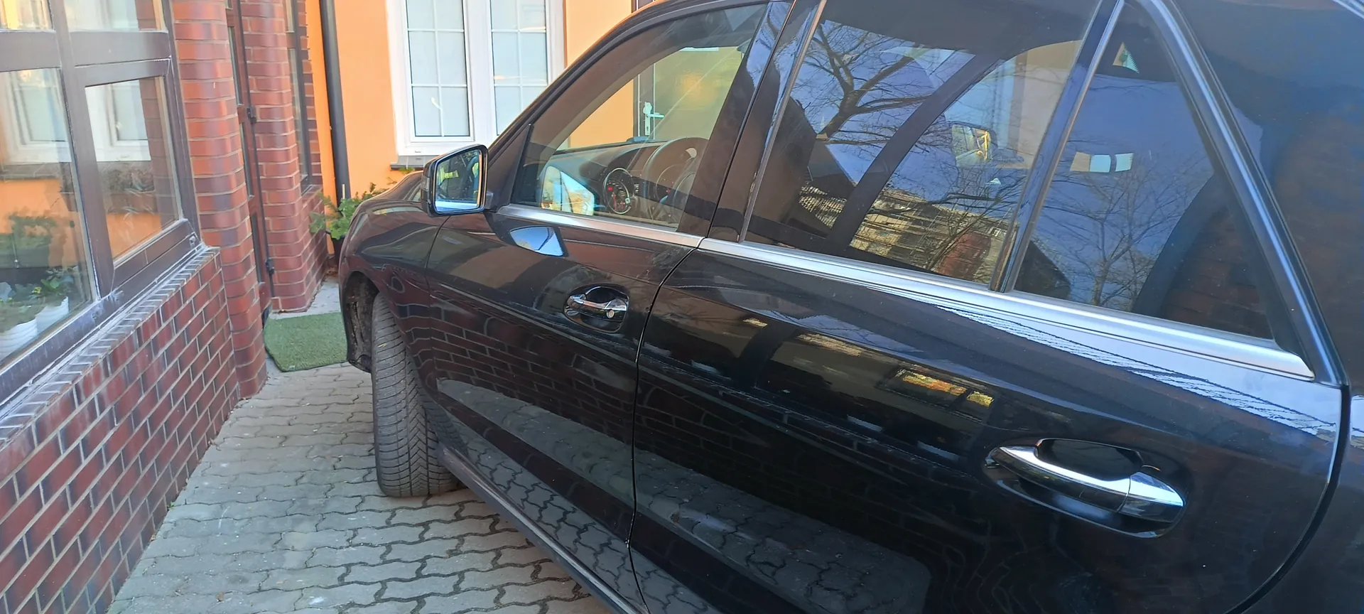 Czarny samochód zaparkowany przy ceglanej ścianie budynku z pomarańczową elewacją i białymi oknami. Widoczne są drzwi samochodu z chromowanymi uchwytami, lusterko boczne oraz odbicie drzew i innych budynków w szybach. Podłoga wyłożona jest kostką brukową, a przy ścianie znajduje się zielony dywanik. W oknach budynku widoczne są doniczki z roślinami.