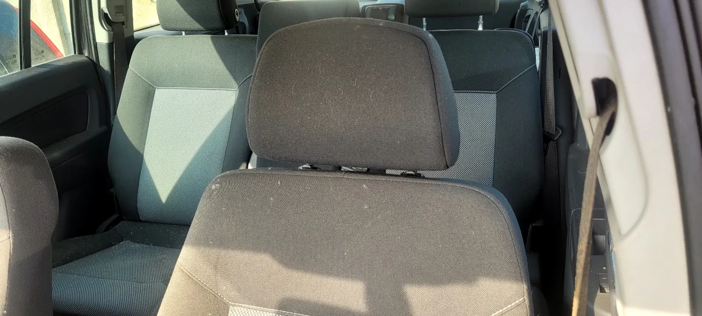 Wnętrze samochodu z widokiem na przednie siedzenia. Siedzenia są ciemnoszare z jasnymi wstawkami na bokach, pokryte kurzem. Widoczne są poduszki głowowe, fragmenty drzwi z oknem po lewej stronie oraz pasy bezpieczeństwa. Światło słoneczne wpada przez okno, rzucając cienie na siedzenia.