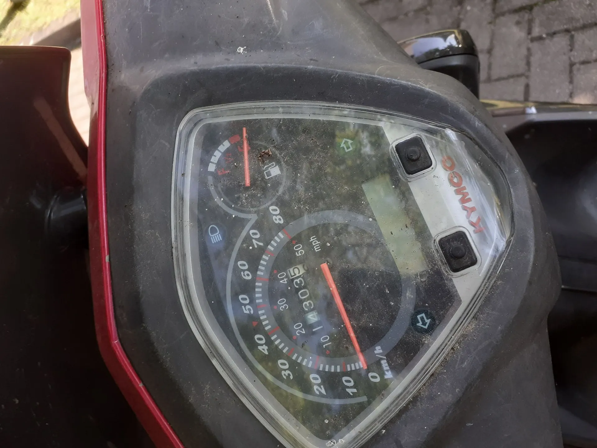Zbliżenie na panel instrumentów skutera marki KYMCO. Widoczny jest prędkościomierz z podziałką w km/h (od 0 do 80) oraz mph, wskaźnik poziomu paliwa, kontrolka świateł oraz kierunkowskazów. Obudowa panelu jest szara i zakurzona, a plastikowa szybka chroniąca wskaźniki jest porysowana i zabrudzona.