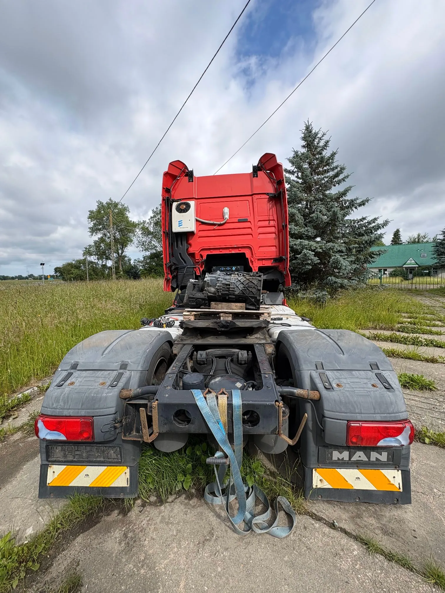 Widok od tyłu na czerwony ciągnik siodłowy marki MAN, zaparkowany na betonowym podjeździe w otoczeniu trawy i drzew. Pojazd nie posiada naczepy, odsłaniając siodło oraz podwozie. Na tylnych błotnikach widoczne są pasy odblaskowe w żółto-pomarańczowe skośne pasy oraz napis 'MAN'. Do ramy pojazdu przymocowany jest niebieski pas transportowy.