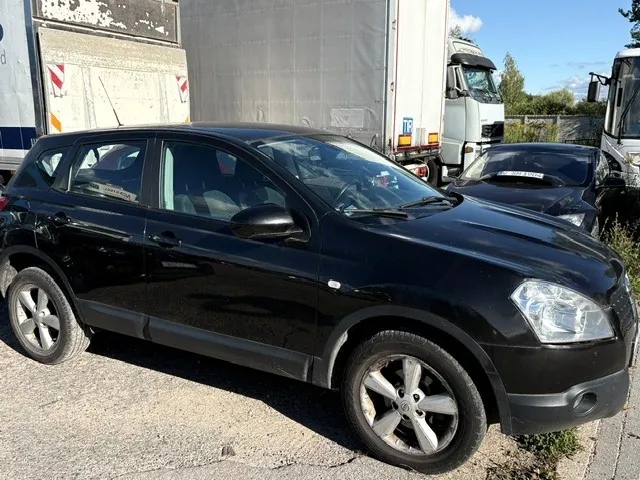 Czarny samochód osobowy typu hatchback zaparkowany na utwardzonym placu. W tle widoczne są białe ciężarówki oraz inny czarny samochód. Niebo jest jasnoniebieskie z kilkoma chmurami.