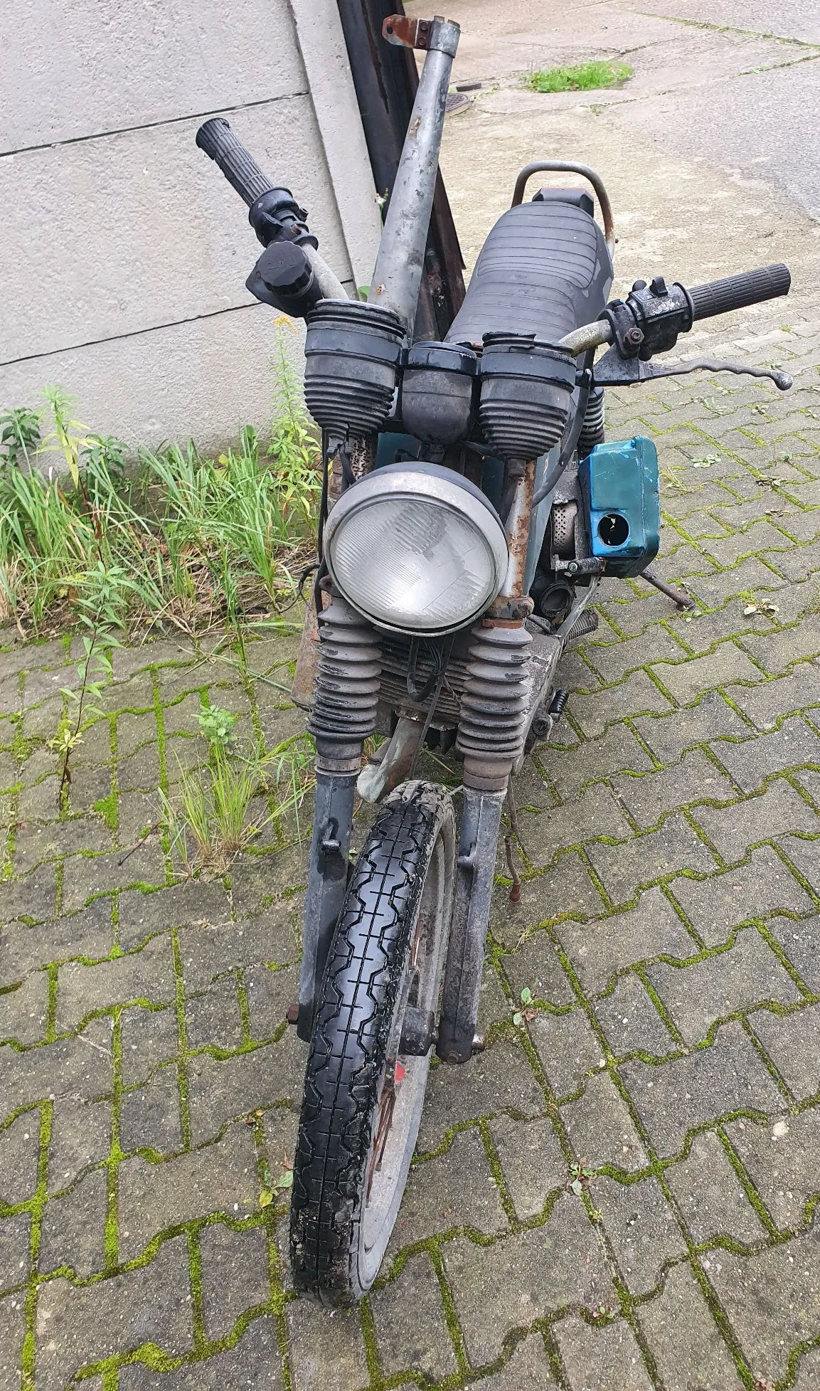 Widok z przodu na stary, zniszczony motocykl zaparkowany na kostce brukowej. Pojazd posiada okrągły reflektor, czarne manetki i siedzenie. Widoczne są ślady rdzy na elementach metalowych oraz zabrudzenia. Po prawej stronie znajduje się niebieski element plastikowy. W tle widać szarą ścianę i kępki trawy wyrastające spomiędzy płyt chodnikowych.