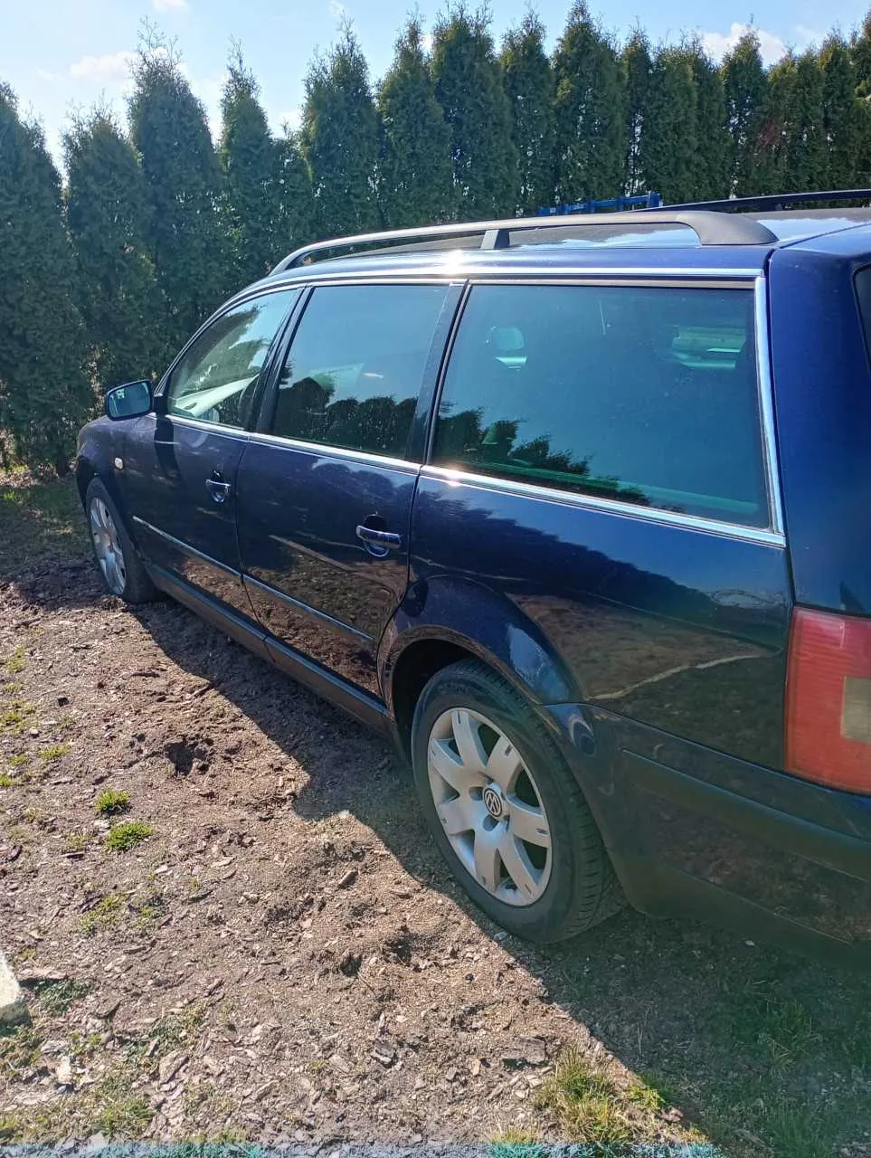 Ciemnoniebieski samochód typu kombi marki Volkswagen zaparkowany na żwirowym terenie z drobnymi kamieniami i rzadką trawą. W tle wysokie, zielone drzewa iglaste oraz jasne niebo z kilkoma chmurami. Samochód posiada dachówkę, srebrne felgi z widocznym logo Volkswagen oraz ciemne szyby. Odbicia na karoserii wskazują na słoneczny dzień.