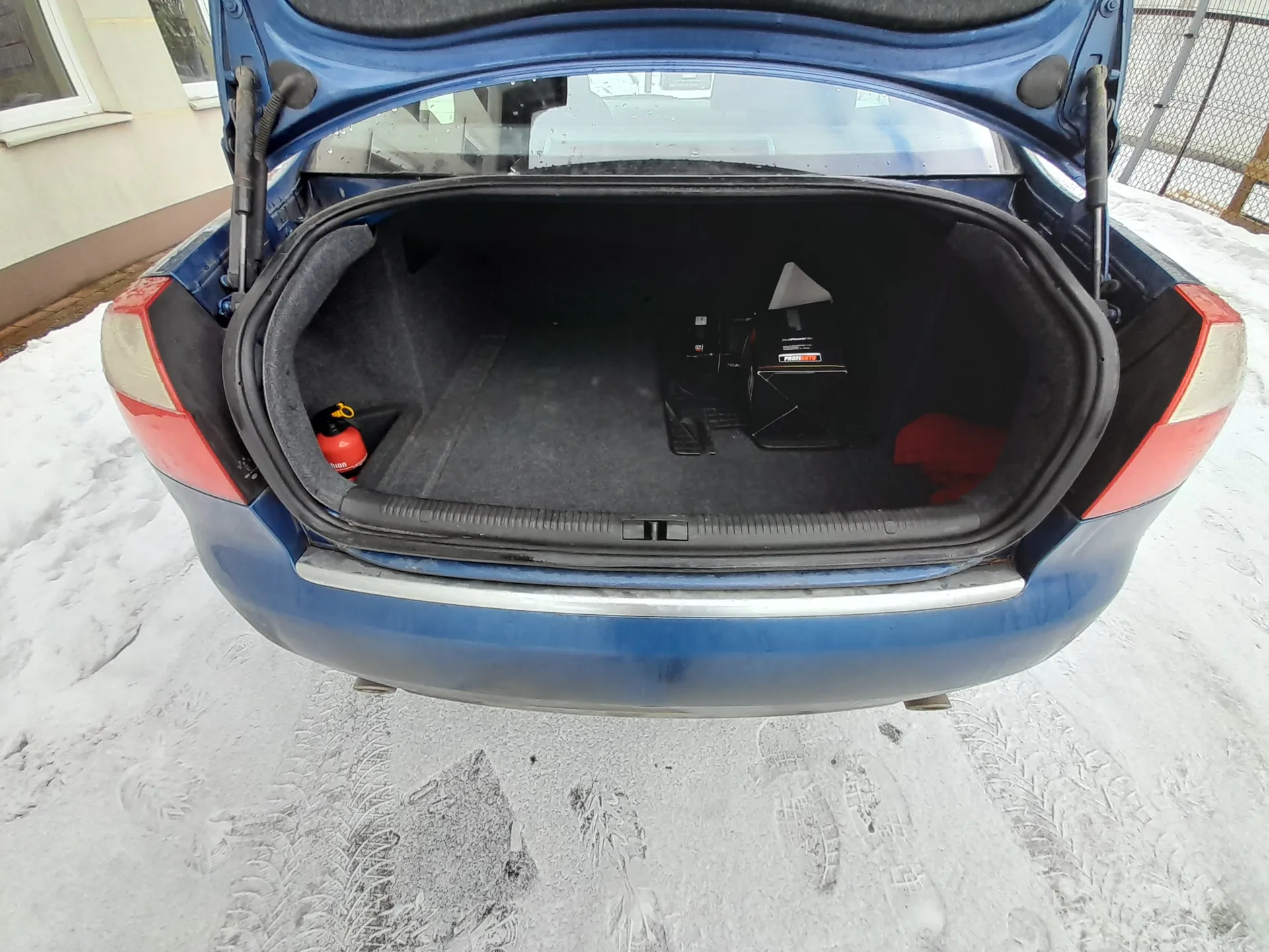 Otwarty bagażnik niebieskiego samochodu zaparkowanego na śnieżnej powierzchni z widocznymi odciskami opon i stóp. Wewnątrz bagażnika znajduje się czerwony gaśnicz, czarne pudełka oraz czerwony przedmiot. W tle widoczne są fragmenty budynku z oknem i metalowego ogrodzenia.