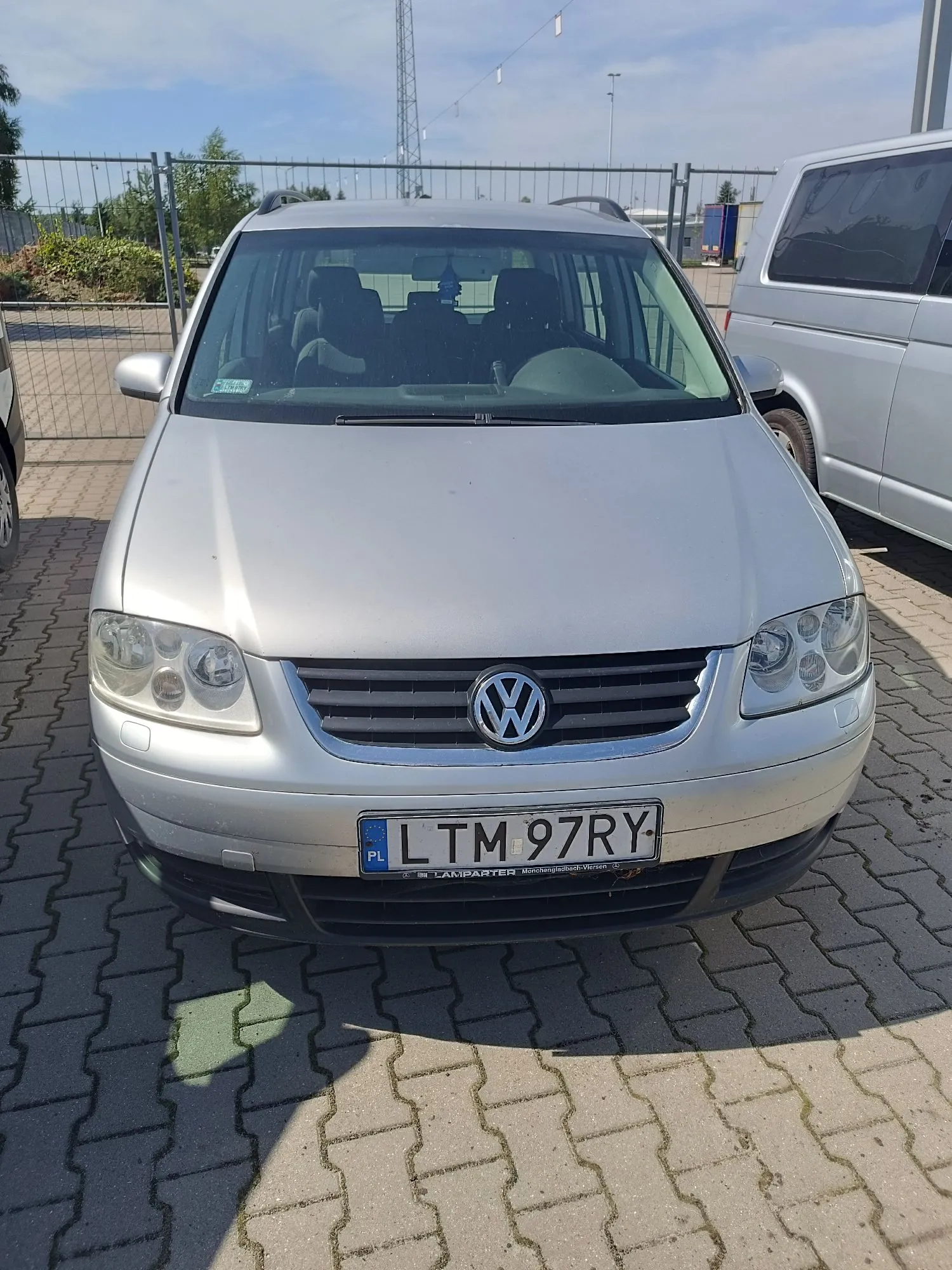 Srebrny samochód marki Volkswagen zaparkowany przodem do widza na kostce brukowej. Na masce i przednim zderzaku widoczne są drobne zarysowania. Samochód posiada polską tablicę rejestracyjną o numerze LTM 97RY. W tle widoczny jest srebrny samochód dostawczy oraz ogrodzenie z siatki.