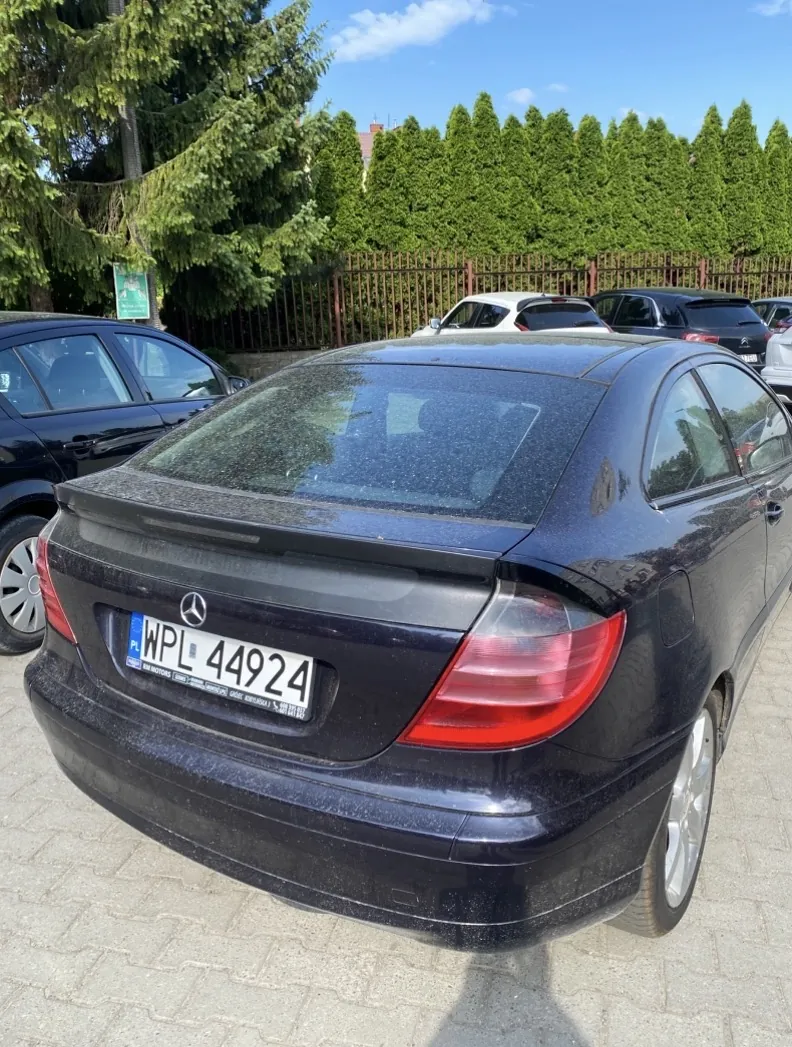 Czarny samochód Mercedes-Benz widziany od tyłu, z tablicą rejestracyjną WPL 44924. Samochód stoi na brukowanej powierzchni, pokryty lekkim kurzem. W tle widoczne są inne pojazdy, metalowe ogrodzenie, gęste zielone drzewa oraz błękitne niebo z kilkoma chmurami. Po lewej stronie znajduje się zielony znak, którego treść nie jest czytelna.