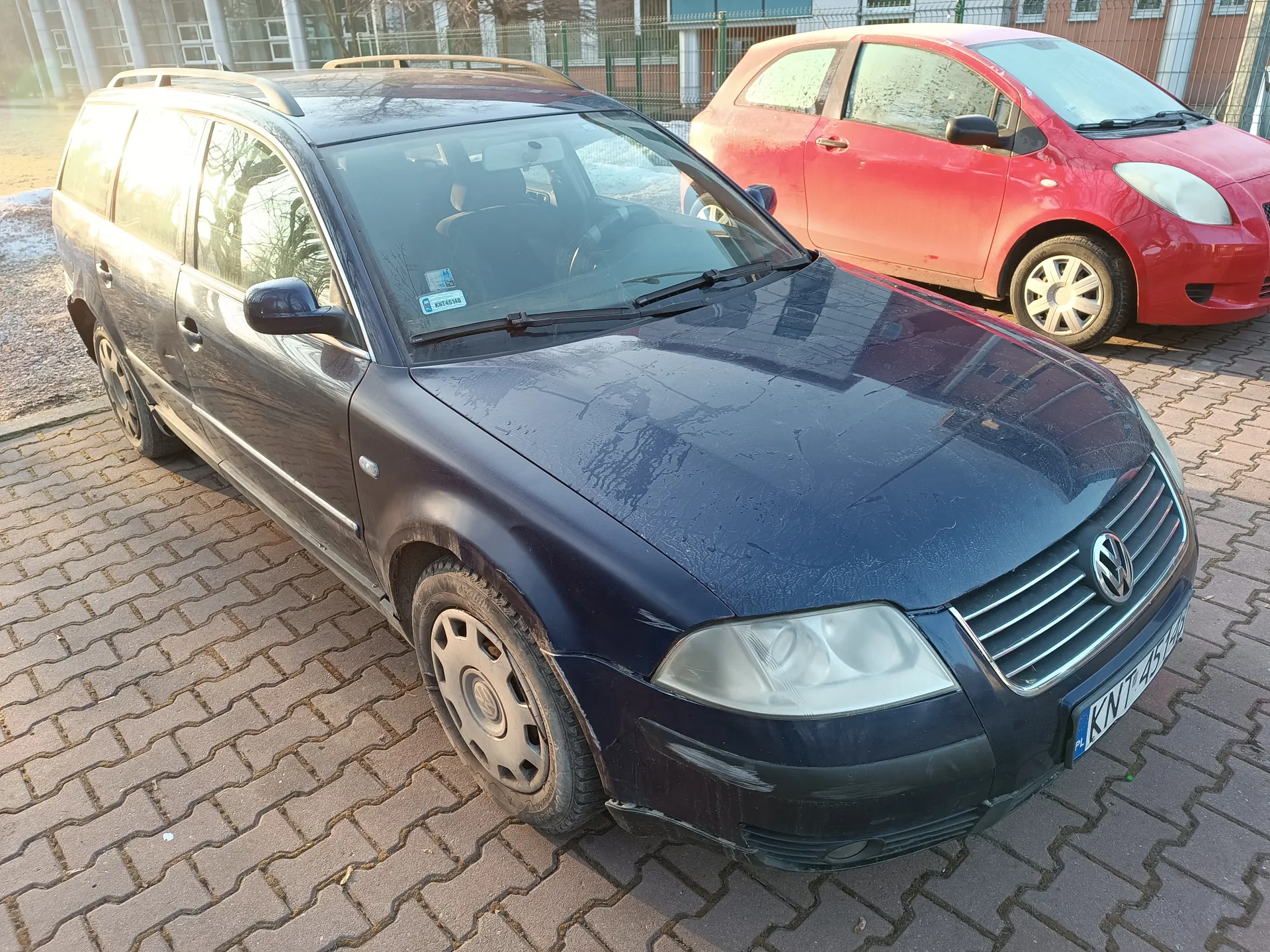 Ciemnoniebieski samochód osobowy typu kombi marki Volkswagen z białą tablicą rejestracyjną KNT 4514 PL, zaparkowany na brukowanym placu. Obok niego czerwony samochód osobowy. W tle budynek z oknami, ogrodzenie i fragmenty śniegu na ziemi. Na przedniej szybie ciemnoniebieskiego samochodu widoczny mały naklejony znaczek.
