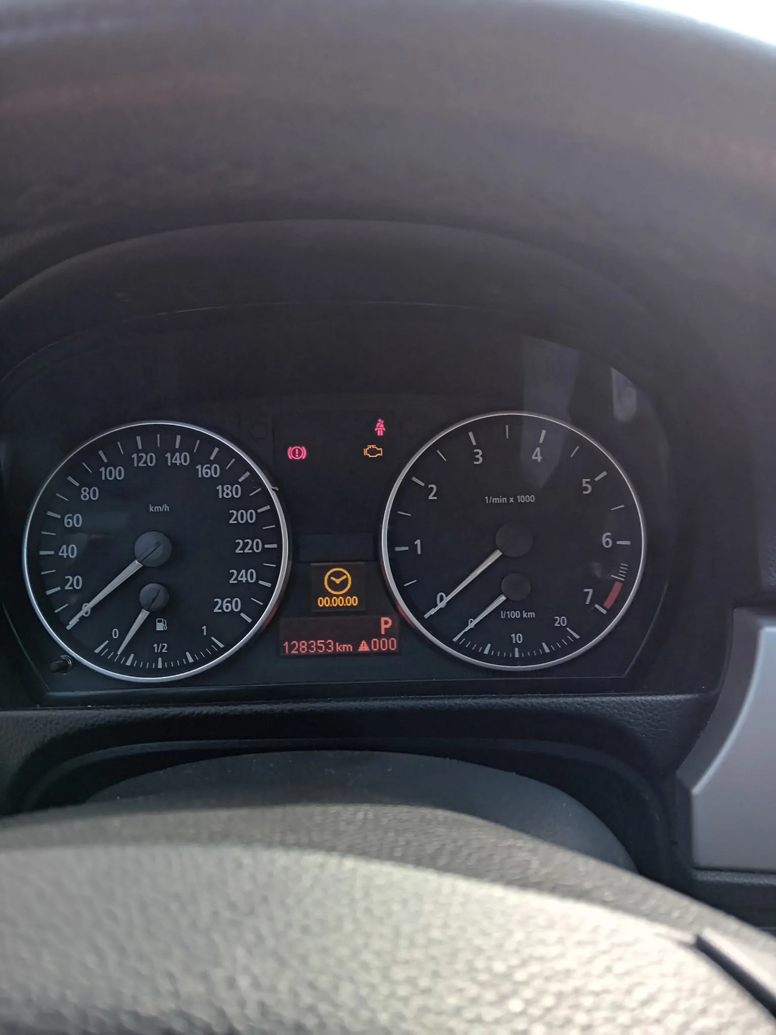 Zbliżenie na deski rozdzielcze samochodu. Po lewej stronie wskaźnik prędkości z zakresem do 260 km/h, obecnie wskazujący 0 km/h, z podwójnym wskaźnikiem paliwa (poziom około 1/2). Po prawej stronie obrotomierz z zakresem do 7 (1/min x 1000), również wskazujący 0. W centrum wyświetlacze cyfrowe: żółty zegar z napisem "00.00.00", czerwony napis "P" oraz przebieg "128353 km" i "000". Widoczne są również lampki ostrzegawcze: czerwona ikona pasów bezpieczeństwa, żółta ikona silnika i czerwona ikona hamulca. Na dole fragment kierownicy.