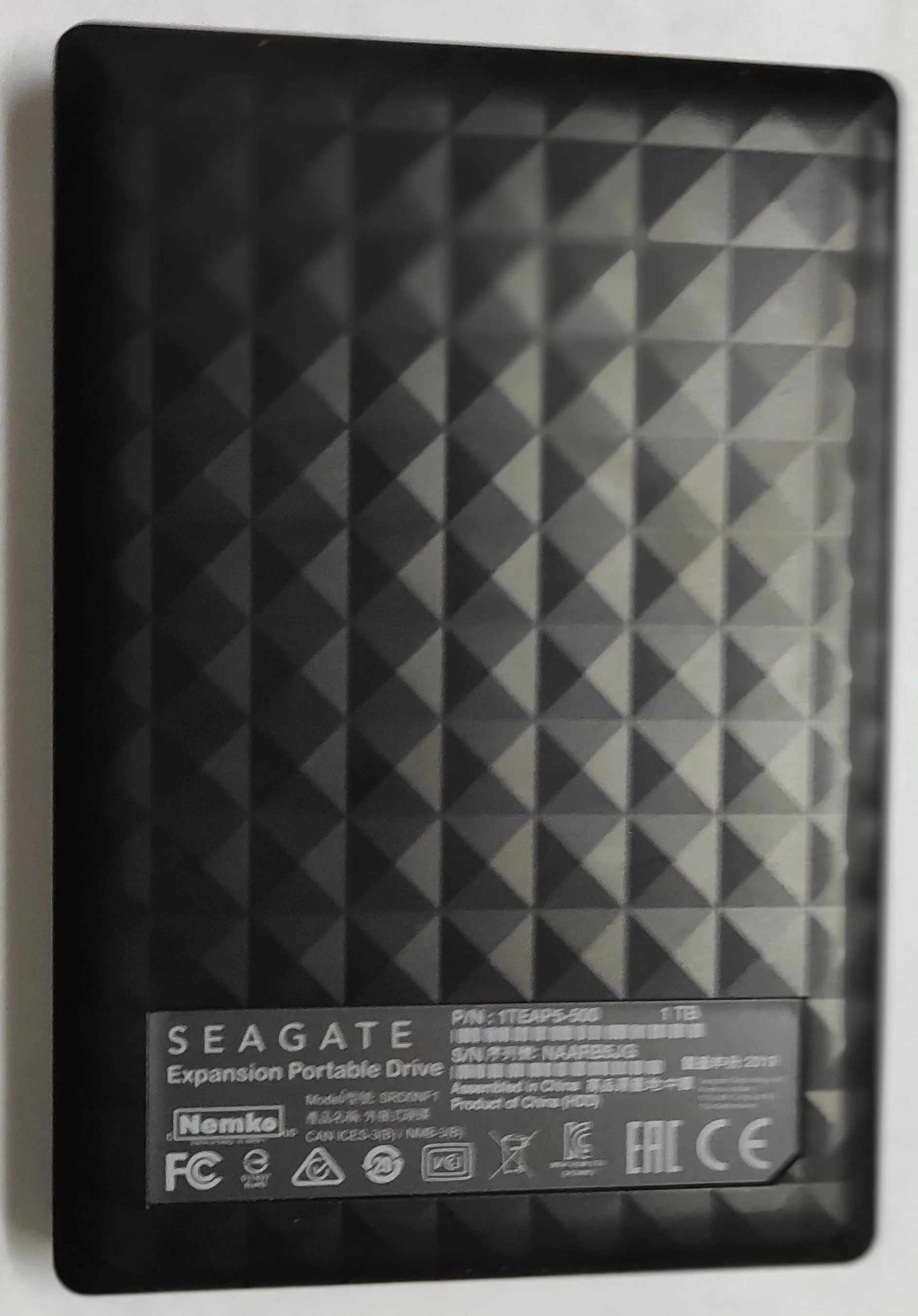 Czarny, prostokątny zewnętrzny dysk twardy marki Seagate Expansion Portable Drive. Obudowa dysku posiada wypukły, geometryczny wzór z powtarzających się trójkątów. Na dole urządzenia znajduje się srebrna etykieta z tekstem: 'SEAGATE Expansion Portable Drive', numerem seryjnym 'S/N: NAARBSZG', pojemnością '1 TB' oraz różnymi certyfikatami i logotypami, takimi jak Nemko, FC, CE i EAC.