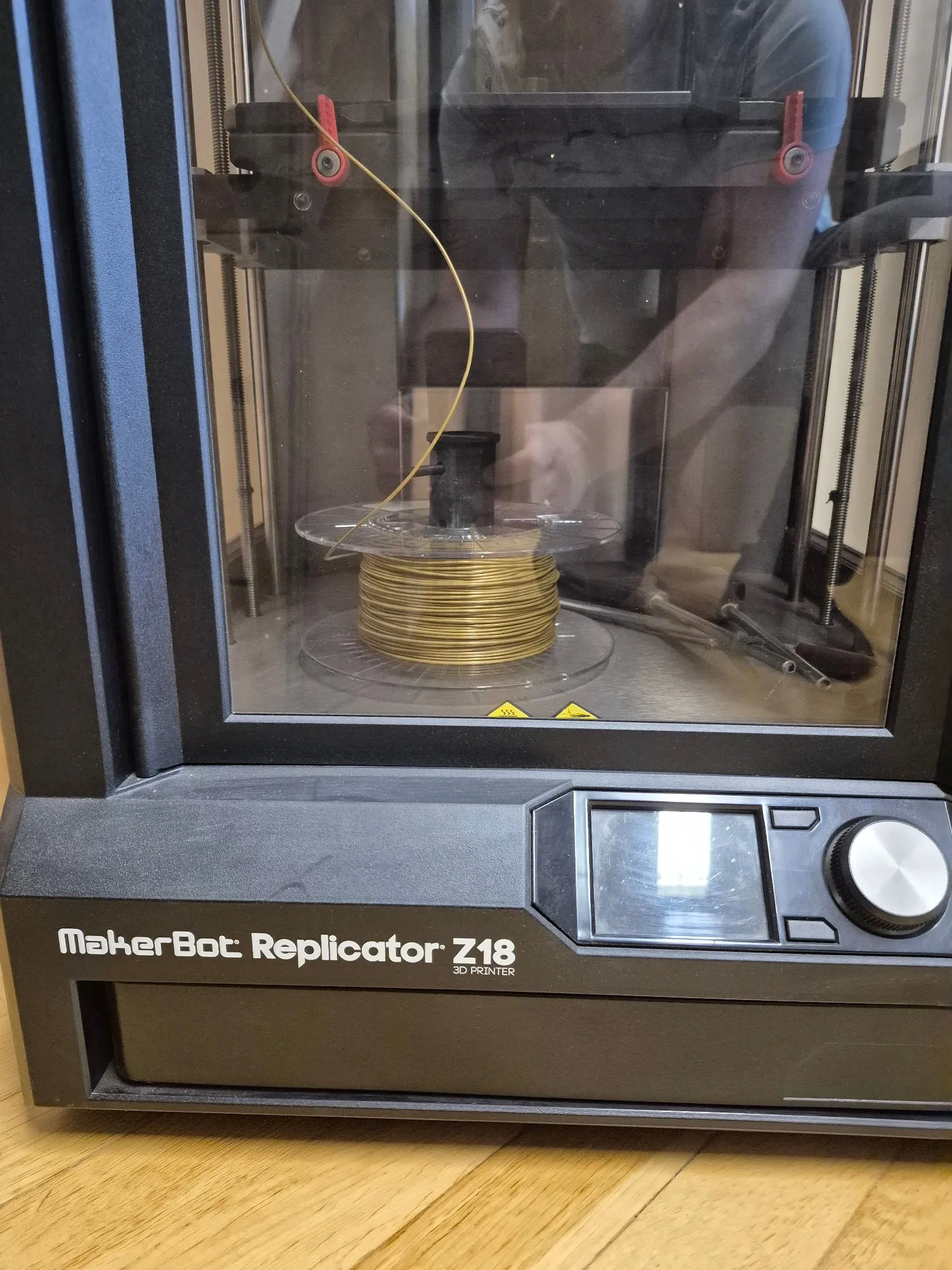 Zdjęcie drukarki 3D MakerBot Replicator Z18. Widoczna jest zamknięta komora z przezroczystą szybą, wewnątrz której znajduje się szpula z żółtym filamentem. Filament prowadzi w górę do głowicy drukującej. Na przednim panelu urządzenia widnieje biały napis 'MakerBot Replicator Z18 3D PRINTER', obok którego znajduje się ekran LCD oraz pokrętło sterujące. Drukarka stoi na drewnianej powierzchni.