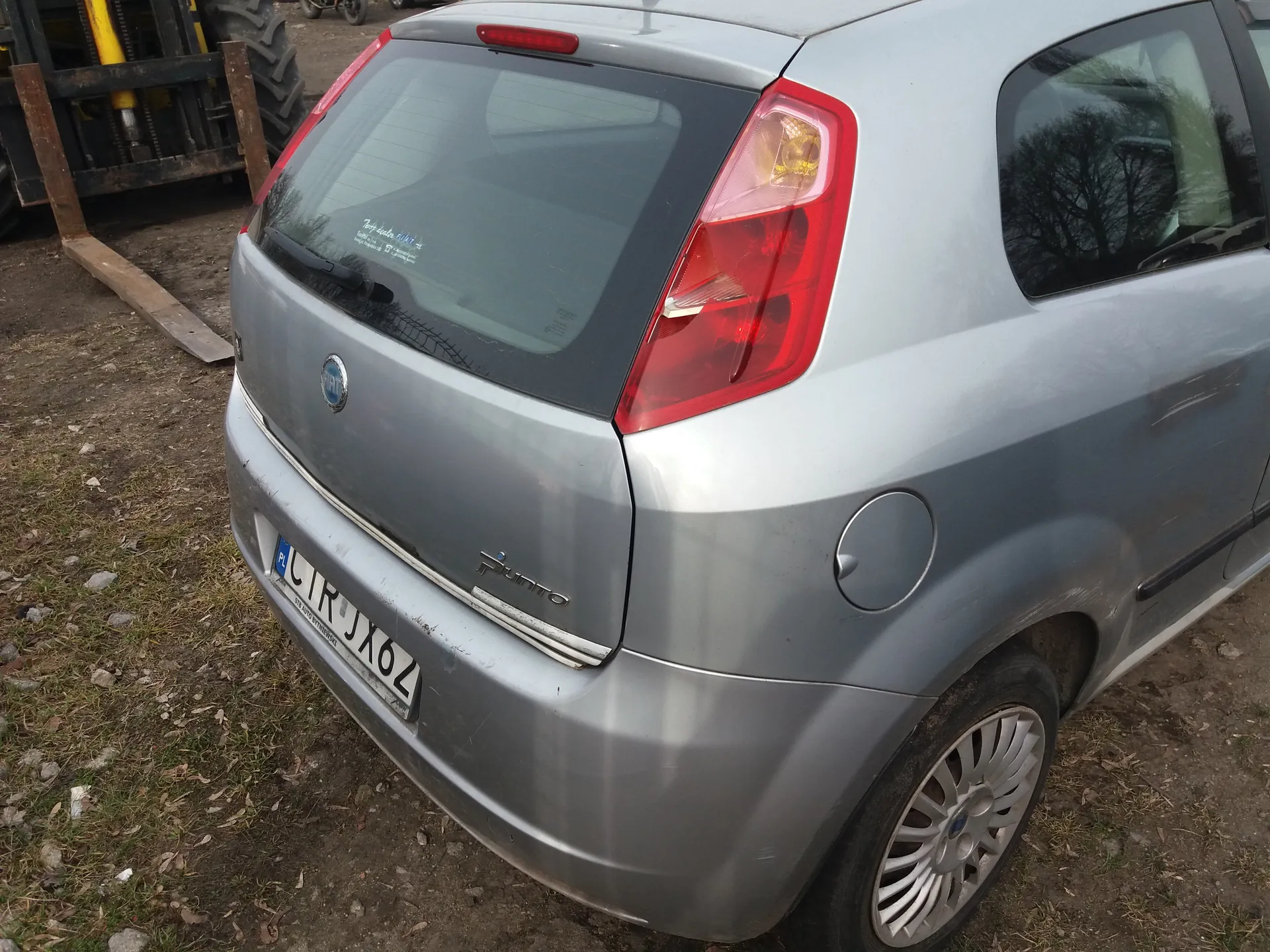 Tył srebrnego samochodu Fiat Punto z widocznym logo marki, napisem "Punto" na bagażniku oraz tablicą rejestracyjną PL CTR JX6Z. W tle fragment maszyny rolniczej z dużym kołem i żółtymi elementami, a także ziemia z trawą i kamieniami. Samochód stoi na nierównym terenie.