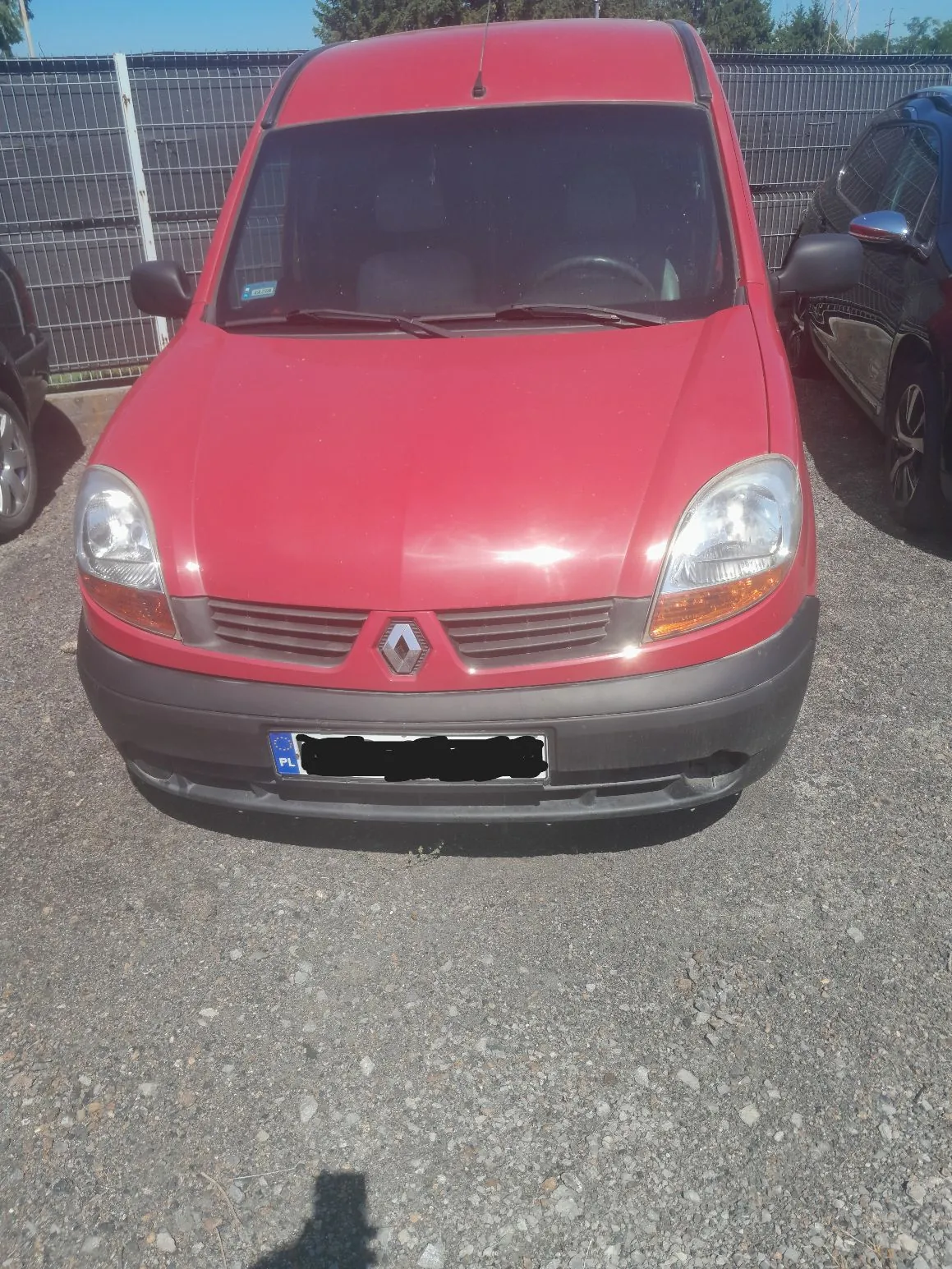 Czerwony samochód marki Renault typu dostawczy, zaparkowany przodem do obiektywu na żwirowym podłożu. Widoczny jest przód pojazdu z charakterystycznym logo rombu na grillu, reflektorami oraz czarnym zderzakiem z tablicą rejestracyjną (zakrytą), z widocznym oznaczeniem kraju PL. W tle znajduje się metalowy płot oraz fragment innego, ciemnego samochodu po prawej stronie.