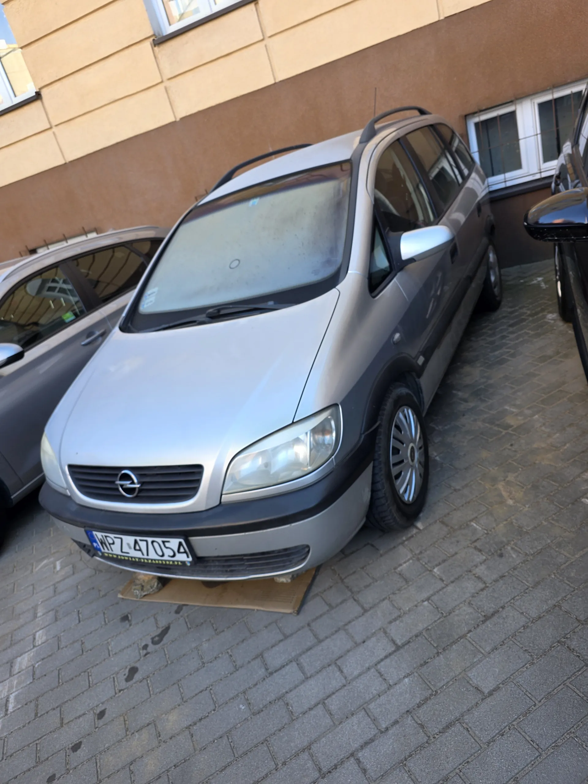 Srebrny samochód osobowy zaparkowany na brukowanej powierzchni. Widoczna tablica rejestracyjna WPL 47054. Po lewej stronie fragment budynku z brązowymi i beżowymi panelami oraz oknami. Na przedniej szybie okrągły znak. Samochód zaparkowany obok innego pojazdu. Obok samochodu leży karton.