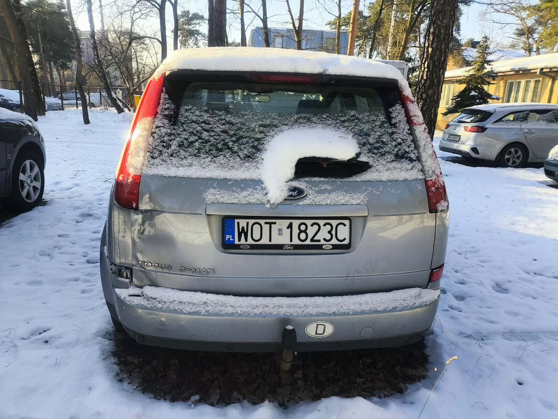 Tył srebrnego samochodu Ford Focus C-MAX pokrytego śniegiem, z widoczną tablicą rejestracyjną WOT 1823C oraz napisem PL po lewej stronie tablicy. Na tle śnieżnego parkingu znajdują się inne pojazdy, drzewa bez liści i budynki. Na tylnym zderzaku widoczny jest napis Focus C-MAX oraz okrągły znak z literą D. Śnieg pokrywa dach, tylną szybę i zderzak pojazdu.