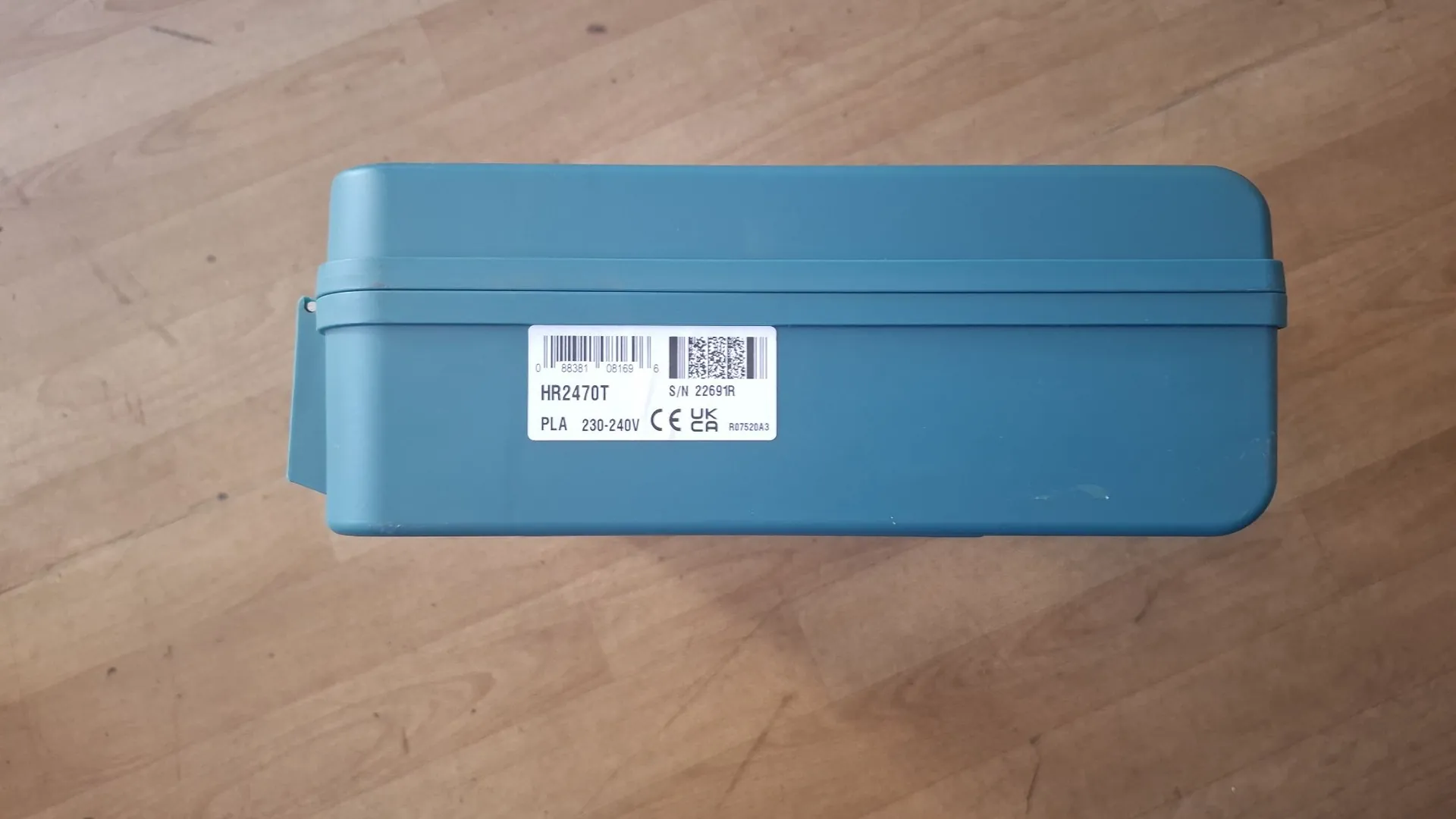 Niebieskie, plastikowe pudełko leżące na drewnianej powierzchni. Na boku pudełka znajduje się biała etykieta z kodem kreskowym, kodem QR oraz tekstem: '0 88381 08169 6', 'S/N 22691R', 'HR2470T', 'PLA 230-240V', 'CE' oraz innymi symbolami certyfikacji.