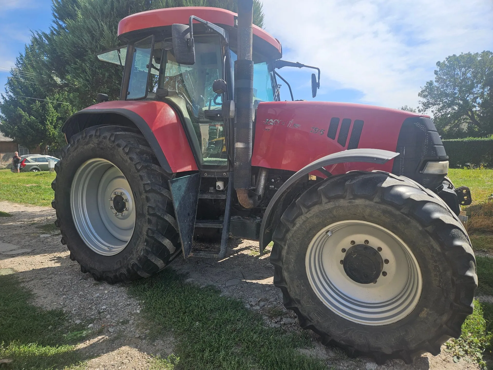 Czerwony traktor rolniczy marki Case IH, model 296, zaparkowany na trawiastym i piaszczystym terenie. Pojazd ma duże, czarne opony z charakterystycznym bieżnikiem i srebrnymi felgami. Widoczna jest przeszklona kabina kierowcy oraz wysoki, czarny wydech. W tle widać zielone drzewa i fragment białego samochodu.