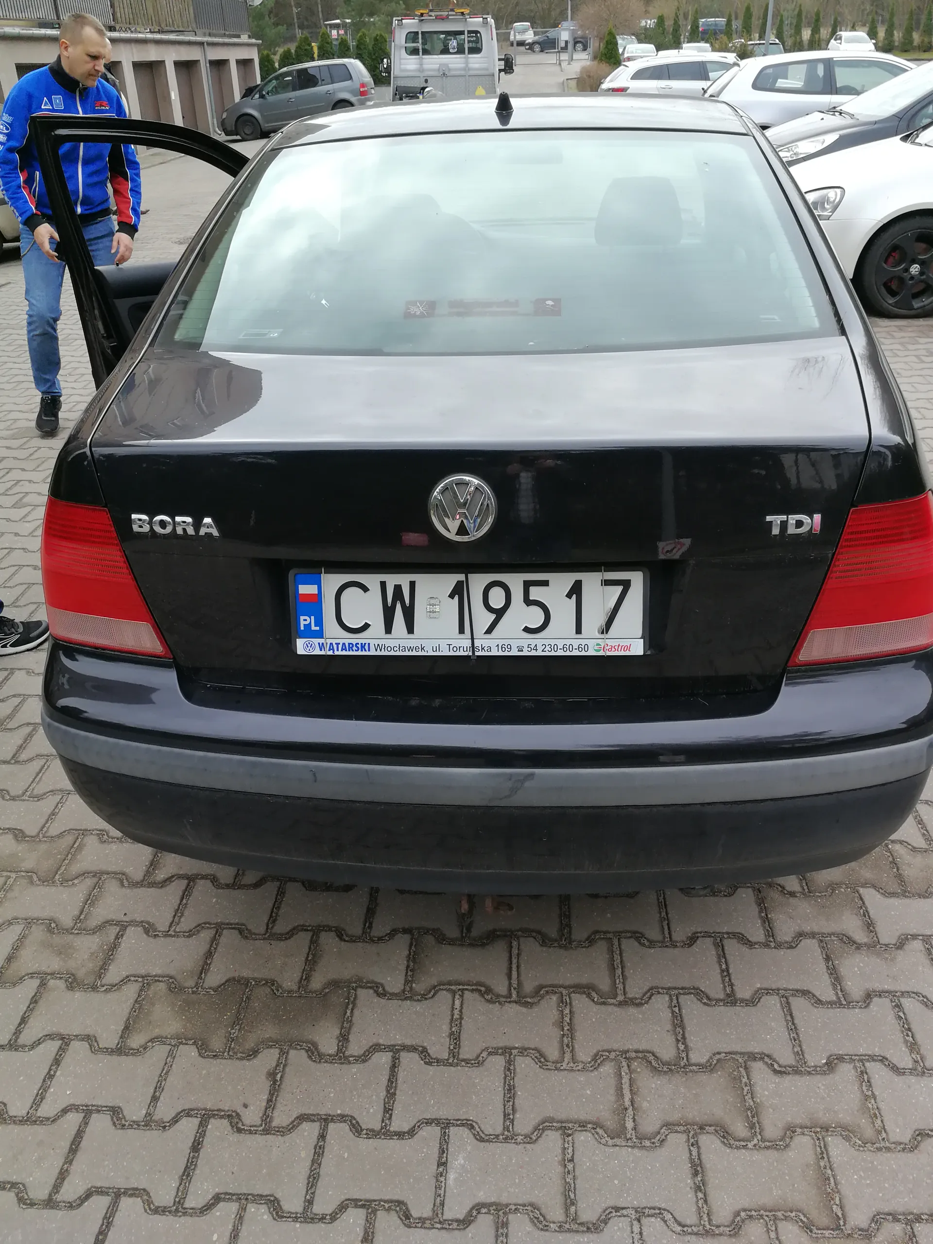 Tył czarnego samochodu Volkswagen Bora TDI z tablicą rejestracyjną CW 19517. Na tablicy widoczny jest polski znak PL oraz naklejka z napisem WATARSKI Włocławek, ul. Toruńska 169 54 230-60-60. Po lewej stronie mężczyzna w niebieskiej kurtce z otwartymi drzwiami kierowcy. W tle inne zaparkowane samochody i budynki. Podłoga wyłożona kostkami brukowymi.