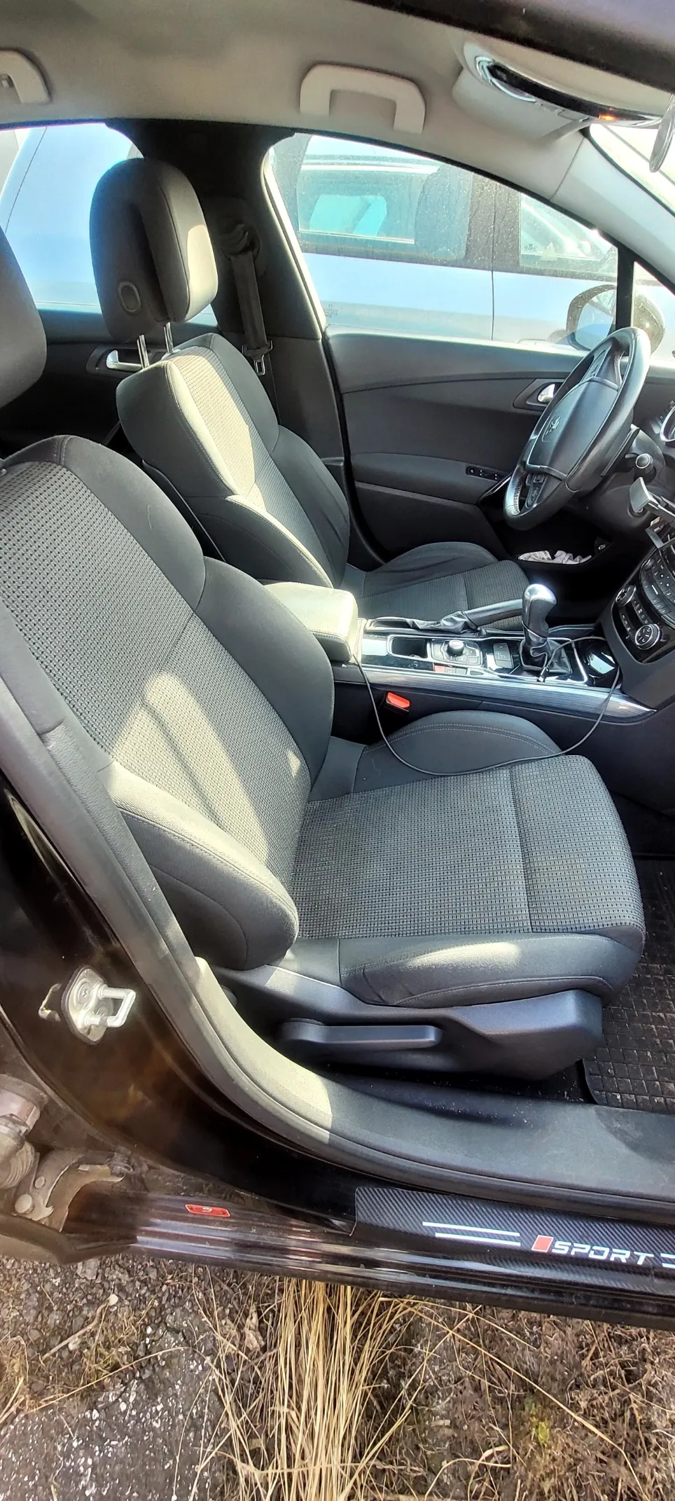 Wnętrze samochodu z widocznymi czarnymi siedzeniami o teksturze siateczkowej, kierownicą po prawej stronie, dźwignią zmiany biegów w środku deski rozdzielczej oraz otwartymi drzwiami z napisem 'SPORT' na progu. W tle widoczne są fragmenty innego pojazdu i zewnętrzne otoczenie z suchą trawą na ziemi.