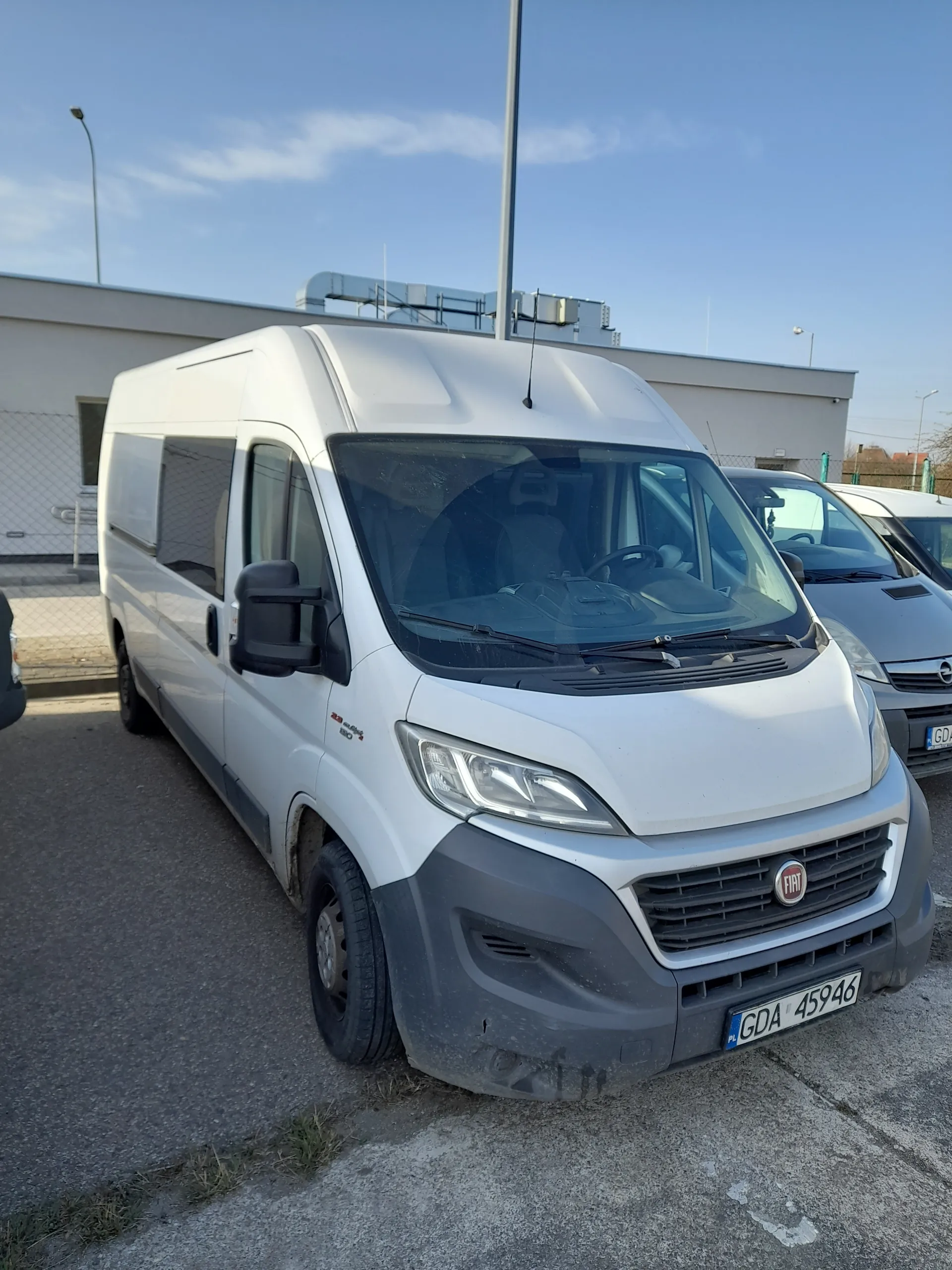 Biały samochód dostawczy marki Fiat zaparkowany na asfaltowej powierzchni. Widoczna tablica rejestracyjna GDA 45946. W tle inne pojazdy, budynek oraz niebo z nielicznymi chmurami. Samochód ma czarny zderzak i logo Fiata na masce.