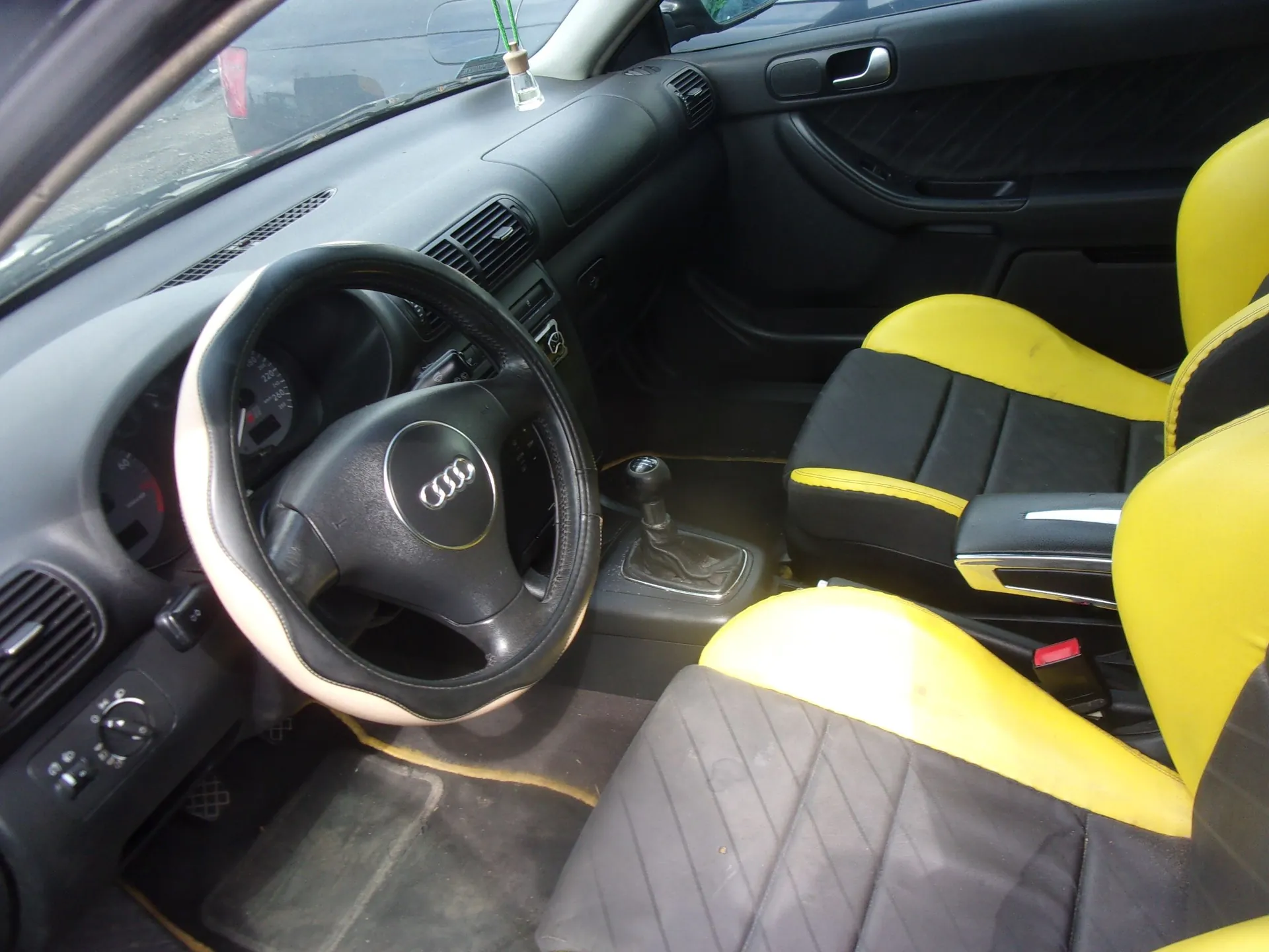 Wnętrze samochodu marki Audi z perspektywy kierowcy. Widoczna jest czarna kierownica z logo Audi na środku, deska rozdzielcza, lewarek zmiany biegów oraz dwa fotele. Fotele są dwukolorowe: czarne z jaskrawożółtymi bokami i zagłówkami. Tapicerka siedzeń ma przeszycia w kształcie rombów.