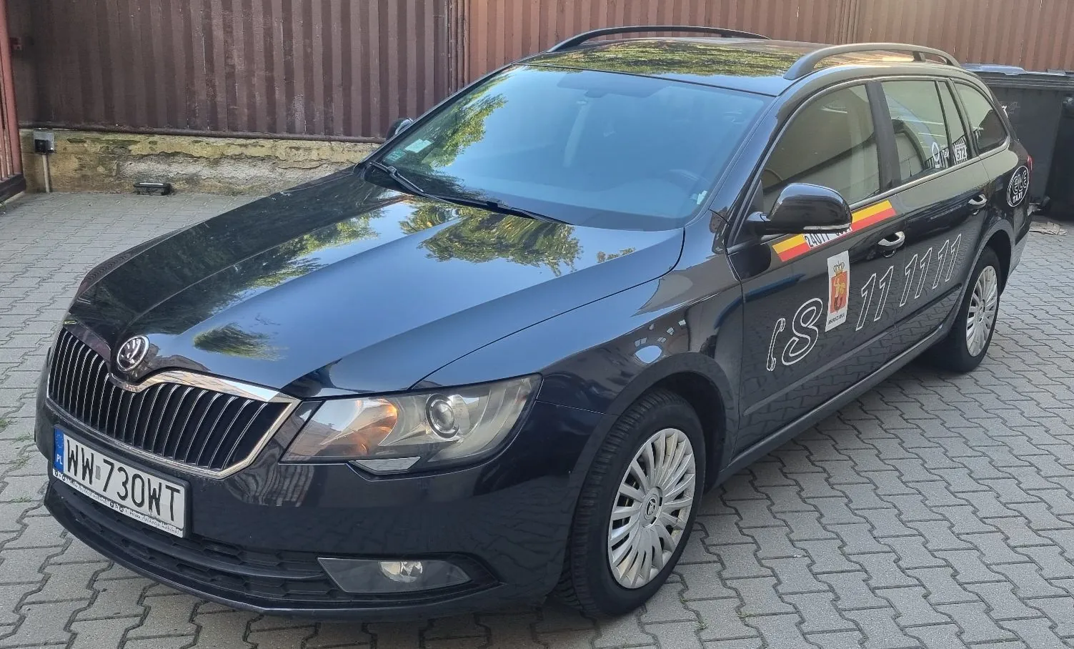 Czarny samochód marki Skoda w nadwoziu kombi zaparkowany na kostce brukowej. Pojazd posiada na boku białe napisy i numer telefonu „(8) 11 11 11”, a nad nimi kolorowy pas w barwach flagi. Tablica rejestracyjna samochodu to WW 730WT.