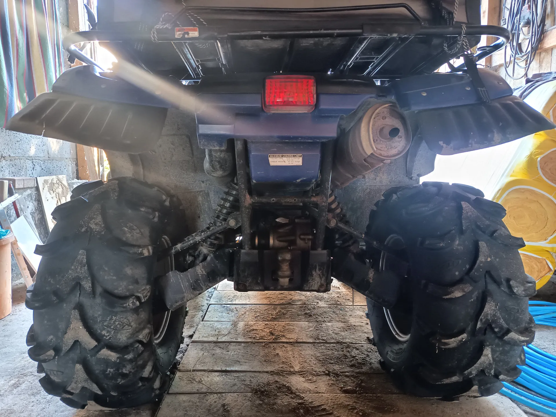 Tylny widok niebieskiego quadra z dużymi, brudnymi oponami. W centrum czerwone światło tylnego reflektora, poniżej tabliczka z napisem "MAXIMUM LOADING LIMIT" oraz dodatkowym, nieczytelnym tekstem. Po prawej stronie widoczny wydech, a w tle betonowa ściana garażu, narzędzia, żółty obiekt oraz niebieskie przewody. Podłoga z drewnianych desek pokryta brudem.