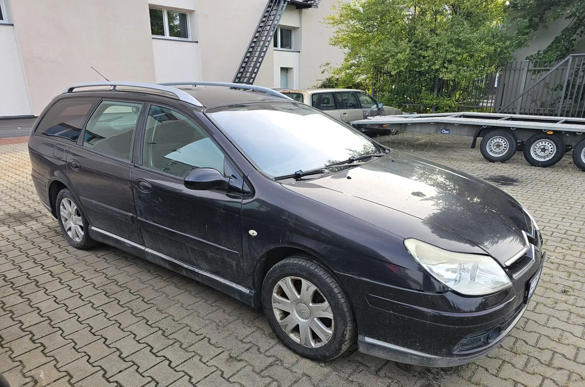 Czarny samochód osobowy typu kombi zaparkowany na kostce brukowej. Pojazd jest zakurzony, posiada srebrne relingi dachowe i aluminiowe felgi. W tle widoczny jest jasny budynek z oknami, schody przeciwpożarowe oraz zaparkowana przyczepa i inny samochód.