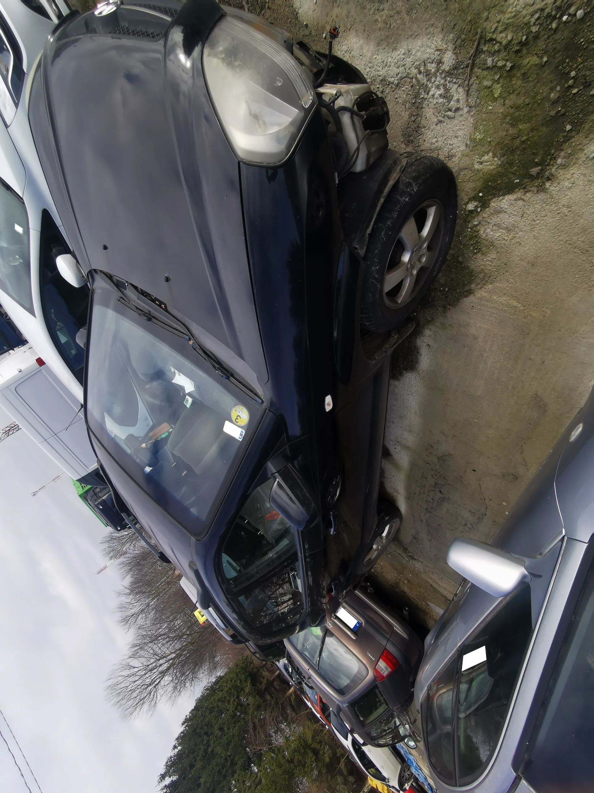 Czarny samochód osobowy z uszkodzonym przodem, zaparkowany na żwirowym placu. Wokół znajdują się inne pojazdy, w tym brązowy i srebrny samochód. Na przedniej szybie czarnego samochodu widoczne są naklejki, w tym żółta z niewyraźnymi symbolami. Tło: drzewa bez liści, pochmurne niebo oraz inne pojazdy w tle.