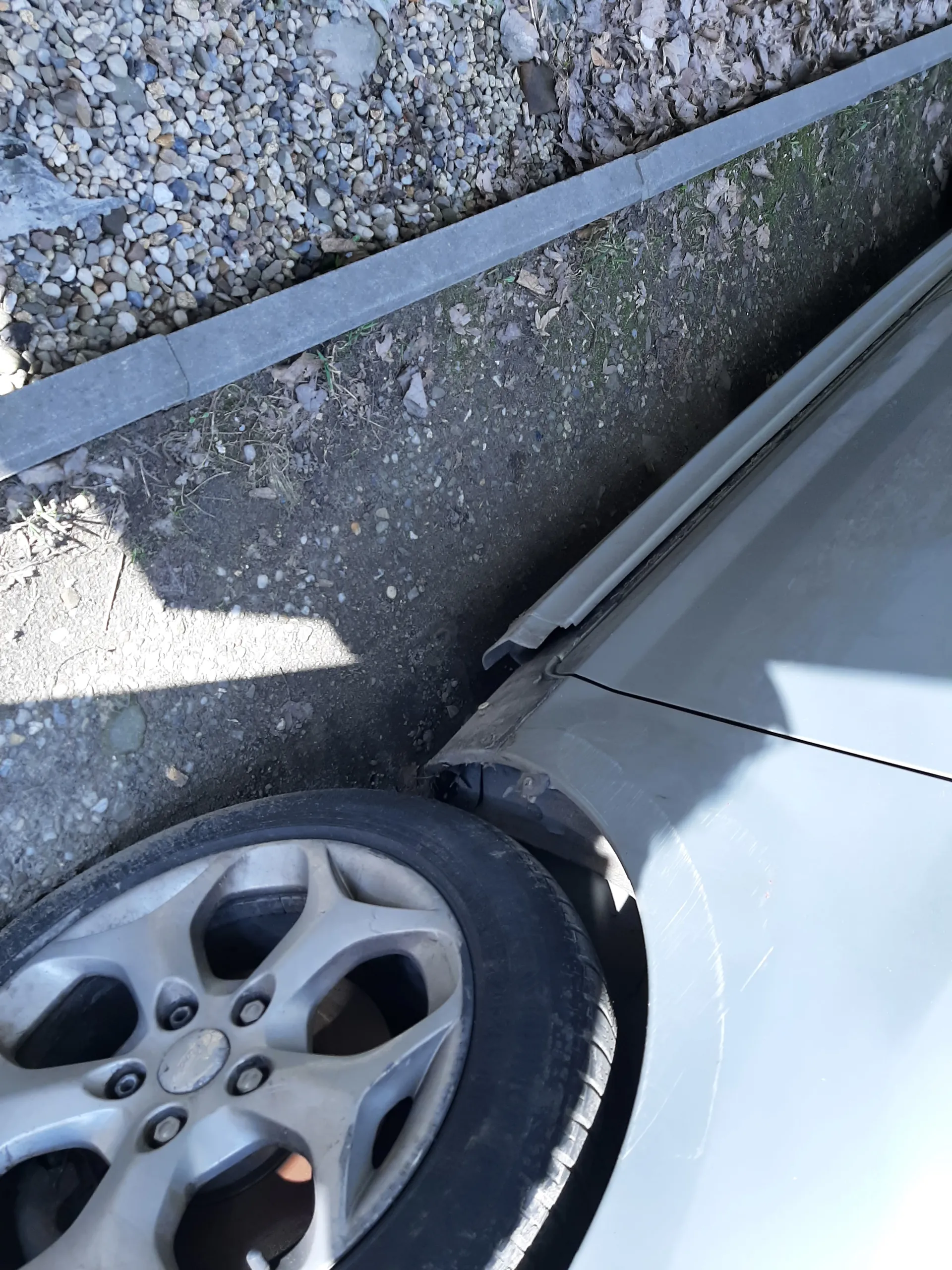 Fragment szarego samochodu z widocznym przednim kołem i dolną częścią karoserii. Koło ma srebrną felgę z pięcioma ramionami i czarną oponą. Pod samochodem znajduje się ziemia z drobnymi kamieniami, suchymi liśćmi i fragmentem szarego chodnika po lewej stronie. Cień samochodu pada na ziemię.