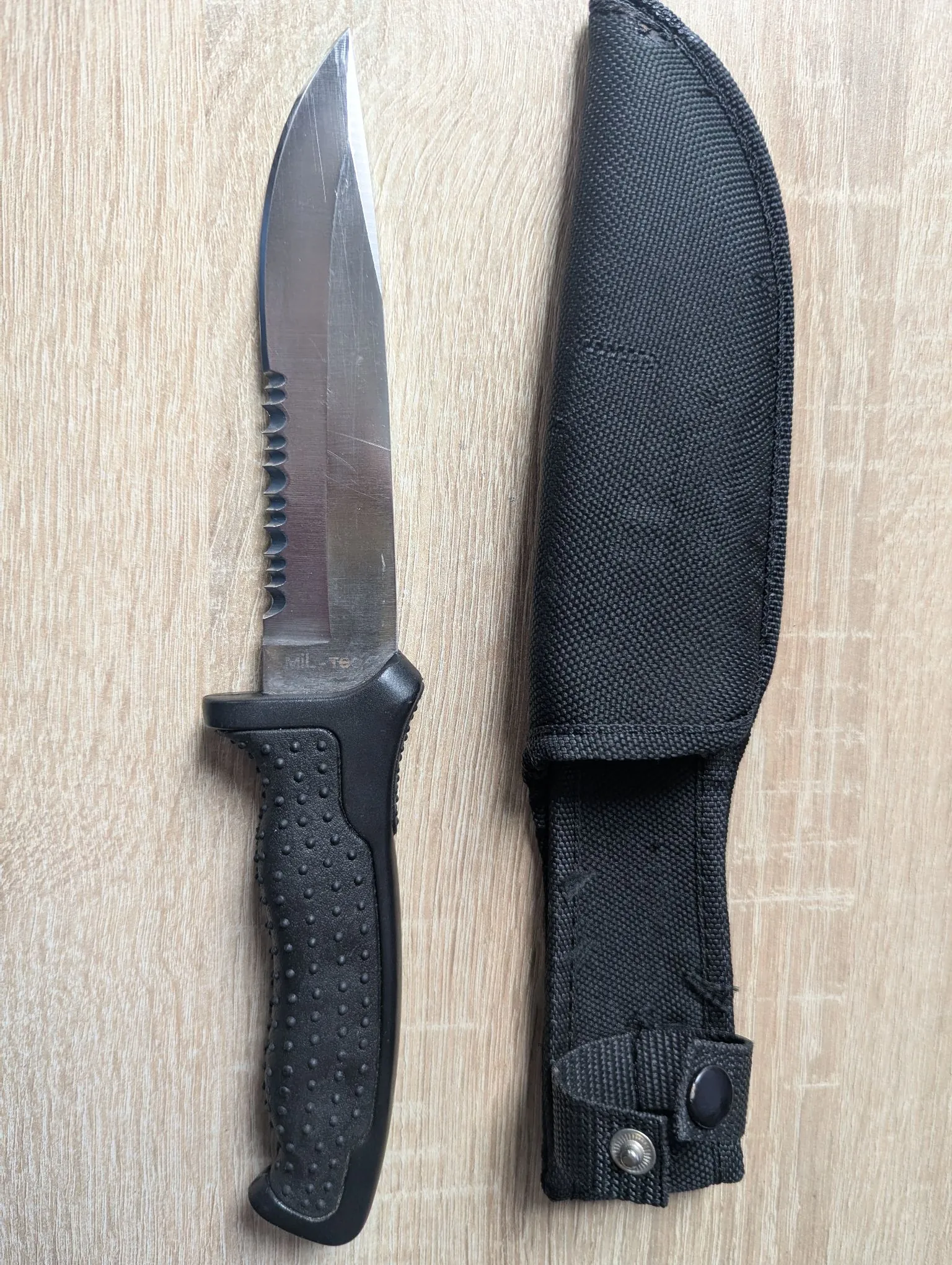 Nóż survivalowy z czarną, teksturowaną rękojeścią i stalowym ostrzem z ząbkowaniem u nasady, obok którego leży czarna, materiałowa pochwa. Na ostrzu widnieje napis 'MIL-TEC'. Przedmioty znajdują się na jasnym, drewnianym tle.