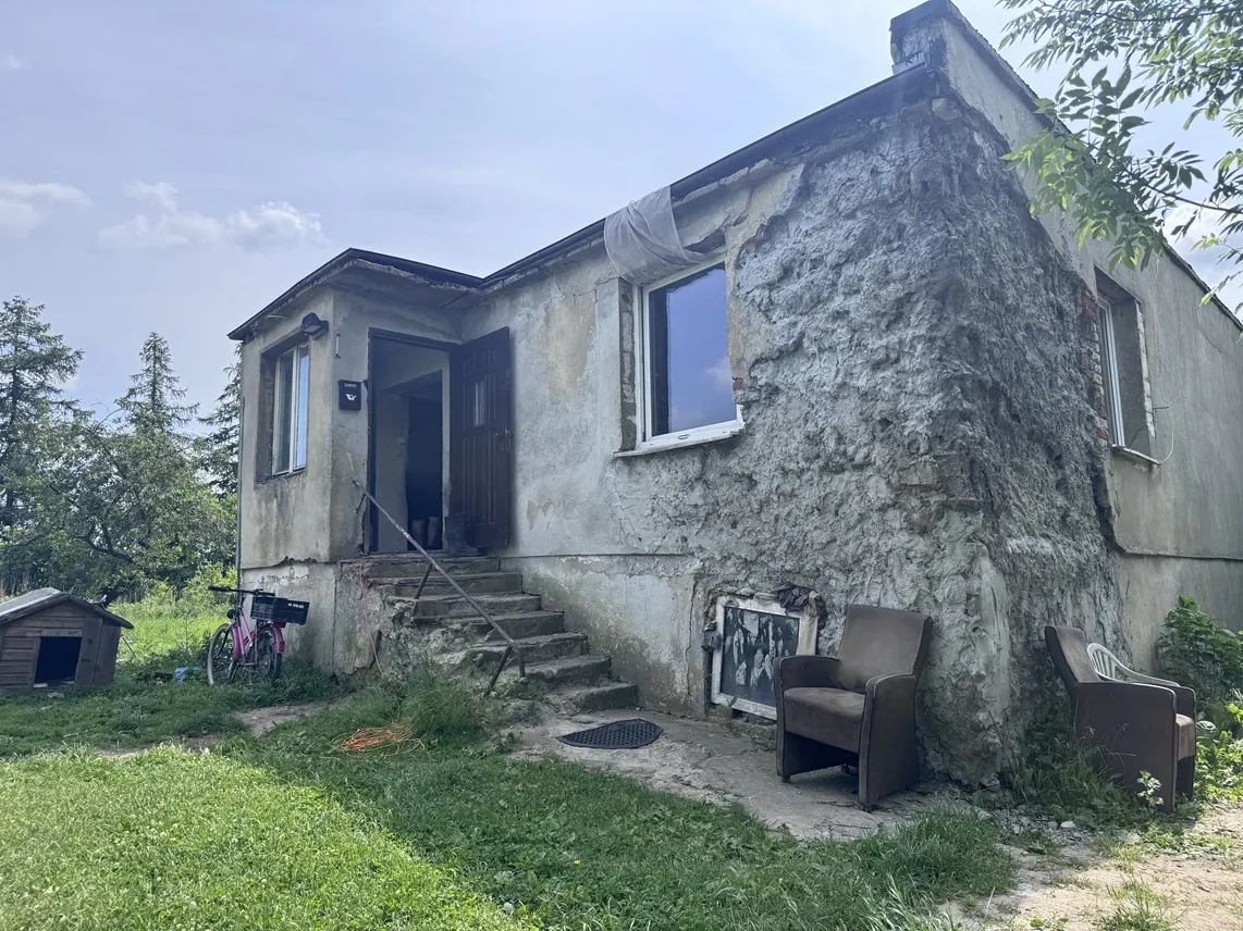 Zdjęcie starego, jednopiętrowego budynku z szarymi, zniszczonymi tynkami. Dom ma otwarte wejście z betonowymi schodami prowadzącymi do wnętrza oraz dwa okna. Przed budynkiem, na trawie, stoją dwa brązowe fotele oraz oparty o ścianę obraz. Po lewej stronie widoczny jest różowy rower oraz mała drewniana buda dla psa. W tle widać zielone drzewa i jasne niebo.