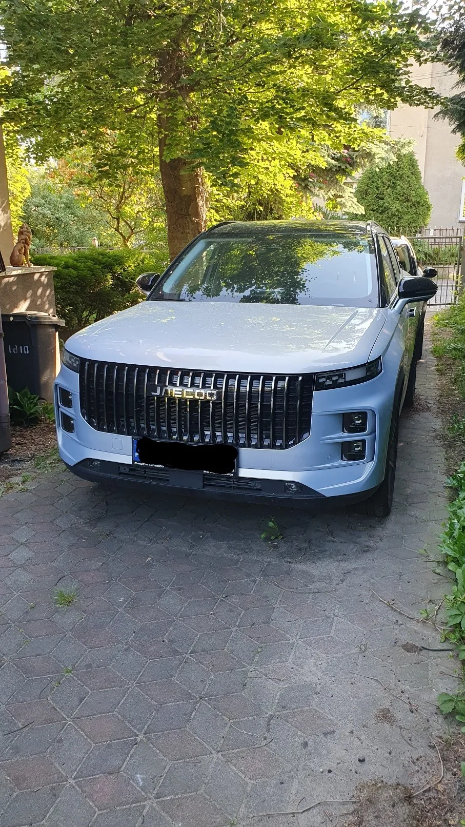 Srebrny samochód marki Li Auto (model L9) zaparkowany na kostce brukowej przed budynkiem. Pojazd ma charakterystyczny szeroki grill z pionowymi żebrami i napisem LI AUTO na środku. W tle widoczne są zielone drzewa i krzewy, a po lewej stronie czarny pojemnik na odpady.