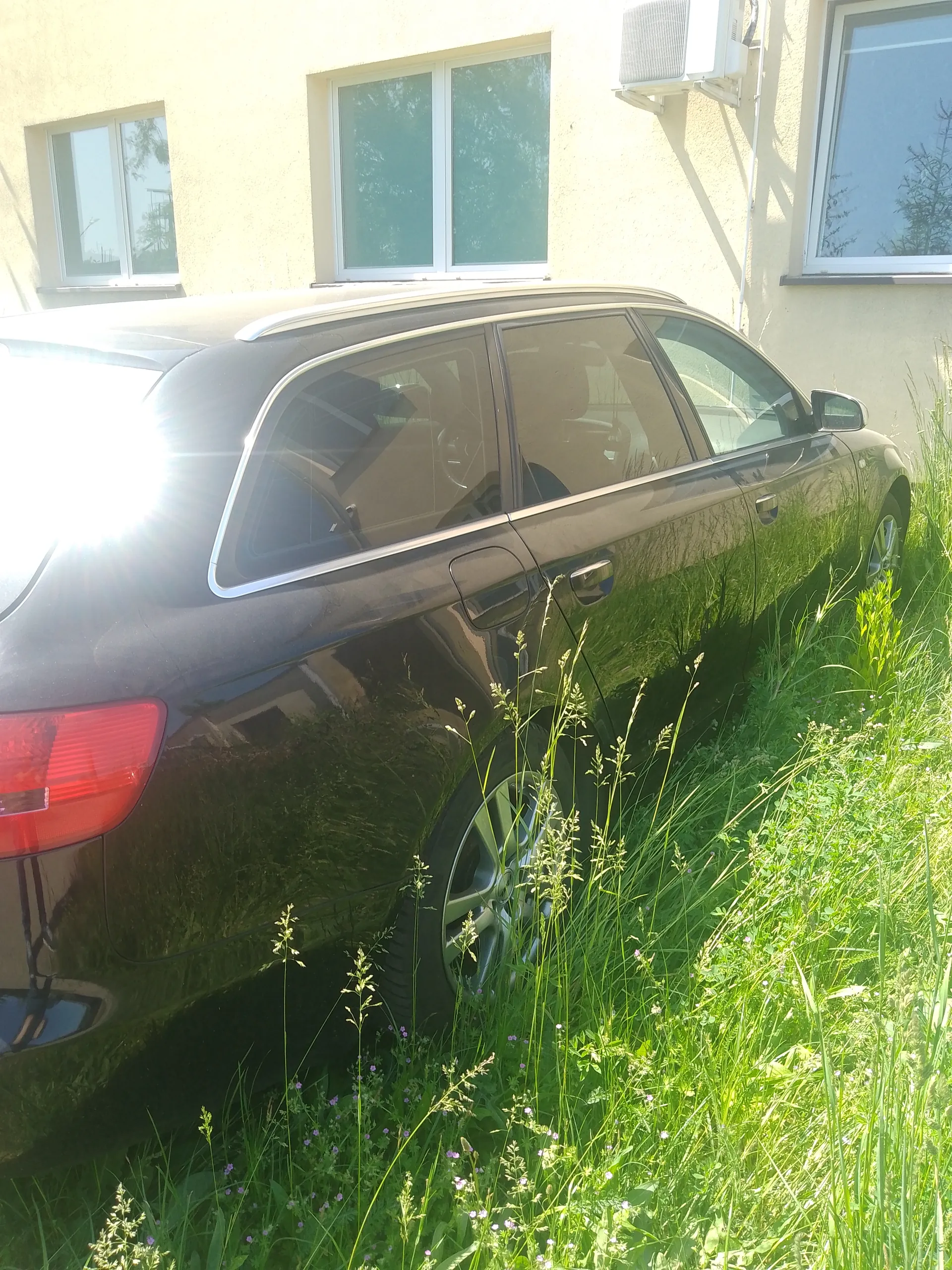 Ciemnoszary samochód typu kombi zaparkowany na wysokiej trawie przed jasnym budynkiem z trzema oknami i jednostką klimatyzacji na ścianie. Słońce oświetla scenę, powodując silne odblaski po lewej stronie. Wysoka trawa z drobnymi fioletowymi kwiatami otacza pojazd.