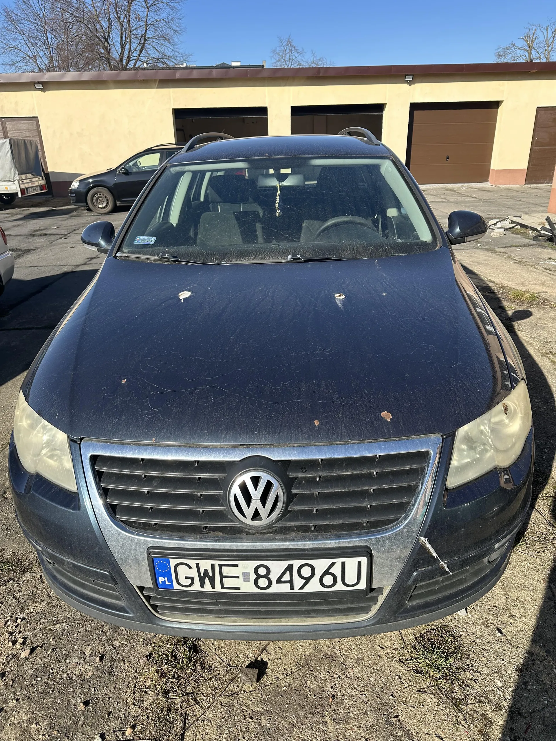 Ciemnoniebieski samochód marki Volkswagen z przodu, z widocznym logo na masce i tablicą rejestracyjną PL GWE 8496U. Na masce znajdują się zadrapania i drobne uszkodzenia. W tle budynek z brązowymi bramami garażowymi, inne pojazdy oraz bezlistne drzewa pod jasnym niebem. Podłoga ziemista z fragmentami trawy.