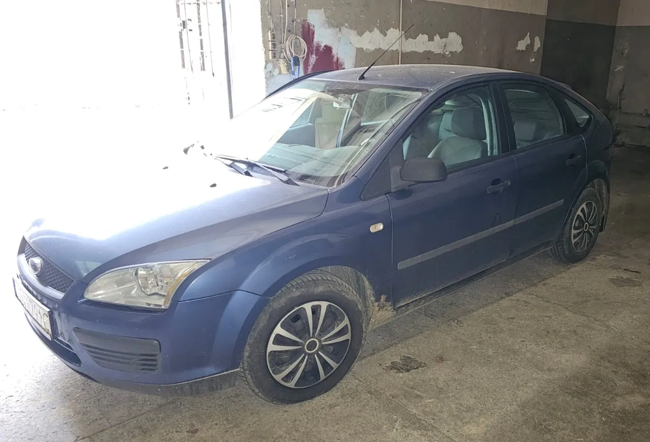 Niebieski samochód osobowy marki Ford Focus zaparkowany wewnątrz garażu lub warsztatu. Pojazd jest zakurzony, widoczne są zabrudzenia na dolnych partiach nadwozia i oponach. Auto stoi pod kątem, ukazując przód oraz prawy bok. W tle widoczna jest jasna otwarta brama oraz ściana z łuszczącą się farbą.