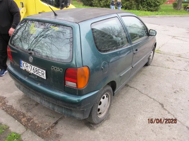 Zielony samochód Volkswagen Polo zaparkowany na betonowej powierzchni. Widoczny tył i bok pojazdu z tablicą rejestracyjną KR 768ER oraz napisem POLO na tylnej szybie. Samochód jest zabrudzony, oznaczenia na kołach i tylnej części. Po lewej stronie stoi osoba w ciemnej odzieży i niebieskich butach. W tle żółty pojazd, zielona trawa, drzewa oraz dwie osoby stojące w oddali. W prawym dolnym rogu obrazu widoczna data 15/04/2026 pomarańczowym kolorem.