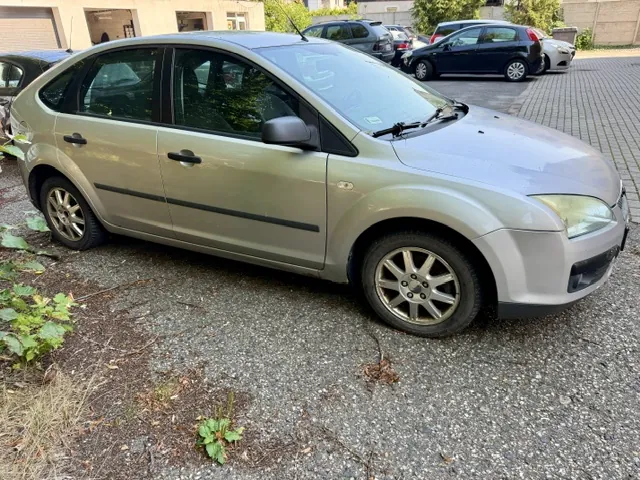 Srebrny samochód osobowy typu hatchback zaparkowany na żwirowym podjeździe. Pojazd jest pokazany z profilu od strony pasażera. W tle widoczne są inne zaparkowane samochody oraz fragmenty budynków.