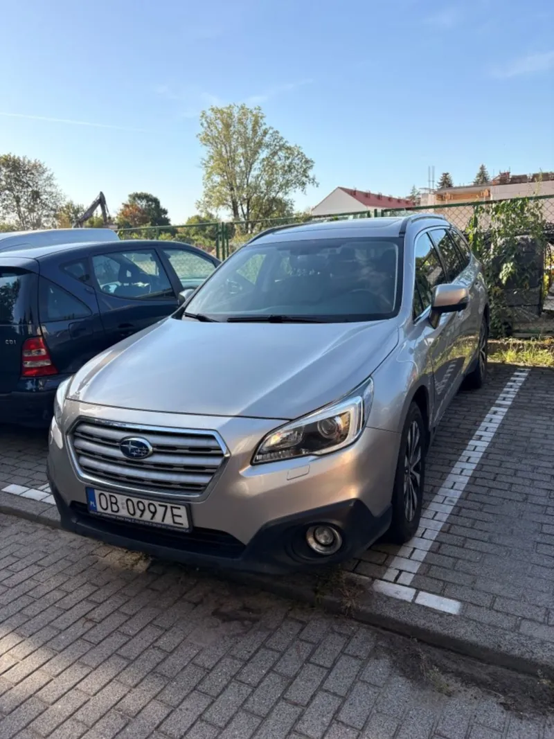 Srebrny samochód marki Subaru Outback zaparkowany na kostce brukowej. Pojazd ma polską tablicę rejestracyjną o numerze OB 0997E. Obok stoi ciemnoniebieski samochód. W tle widoczne są zielone drzewa, ogrodzenie oraz fragmenty budynków pod jasnym, niebieskim niebem.