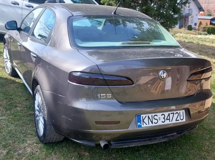 Tylny widok brązowego samochodu marki Alfa Romeo model 159 zaparkowanego na trawie. Na klapie bagażnika widoczne jest logo marki oraz oznaczenie modelu '159'. Pojazd posiada polską tablicę rejestracyjną o numerze KNS 3472U.