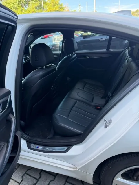 Widok do wnętrza białego samochodu przez otwarte tylne drzwi. W środku znajdują się czarne, skórzane fotele z zagłówkami. Na progu samochodu widoczne jest logo BMW M. Samochód stoi na kostce brukowej.