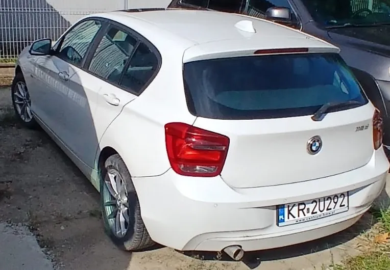 Biały samochód marki BMW w nadwoziu hatchback, zaparkowany tyłem do obiektywu. Widoczna jest tylna część pojazdu z logo BMW, prawą lampą oraz tablicą rejestracyjną o numerze KR 2U292. Samochód ma srebrne felgi aluminiowe i jedną rurę wydechową po lewej stronie.