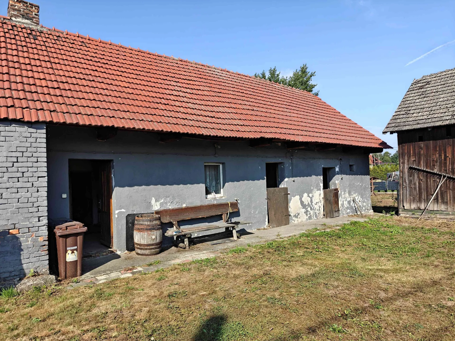 Zdjęcie starego, parterowego budynku z czerwonym, dachówkowym dachem. Ściana budynku jest szara, z widocznymi ubytkami tynku, a lewa część wykonana jest z szarej cegły. Przed budynkiem znajduje się drewniana ławka, drewniana beczka oraz brązowy kosz na śmieci. Po prawej stronie widoczny jest fragment innego, drewnianego budynku. Przed budynkami rozciąga się trawnik pod bezchmurnym, niebieskim niebem.