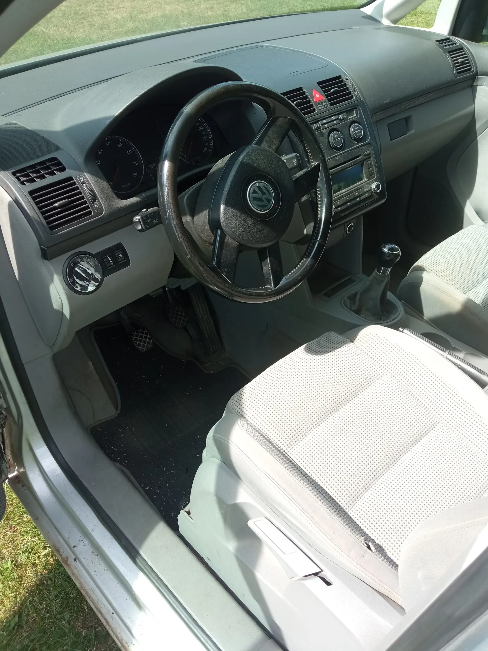 Wnętrze samochodu marki Volkswagen widziane z perspektywy otwartych drzwi kierowcy. Widoczna jest szara deska rozdzielcza, czarna kierownica z logo VW, zegary, radio oraz jasnoszara tapicerka fotela kierowcy.