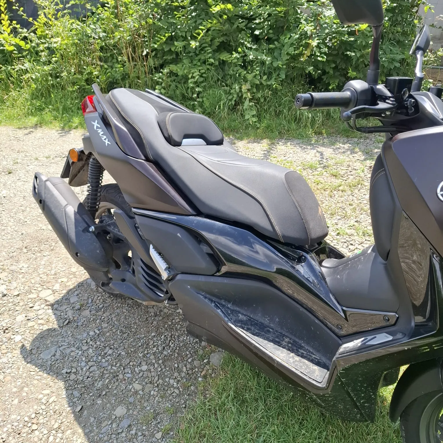 Zdjęcie przedstawiające boczny widok czarnego skutera marki Yamaha z napisem 'Xmax' na tylnej owiewce. Pojazd stoi na żwirowej drodze na tle zielonych krzewów i trawy. Widoczne są detale takie jak czarne siedzenie, rura wydechowa oraz kierownica.