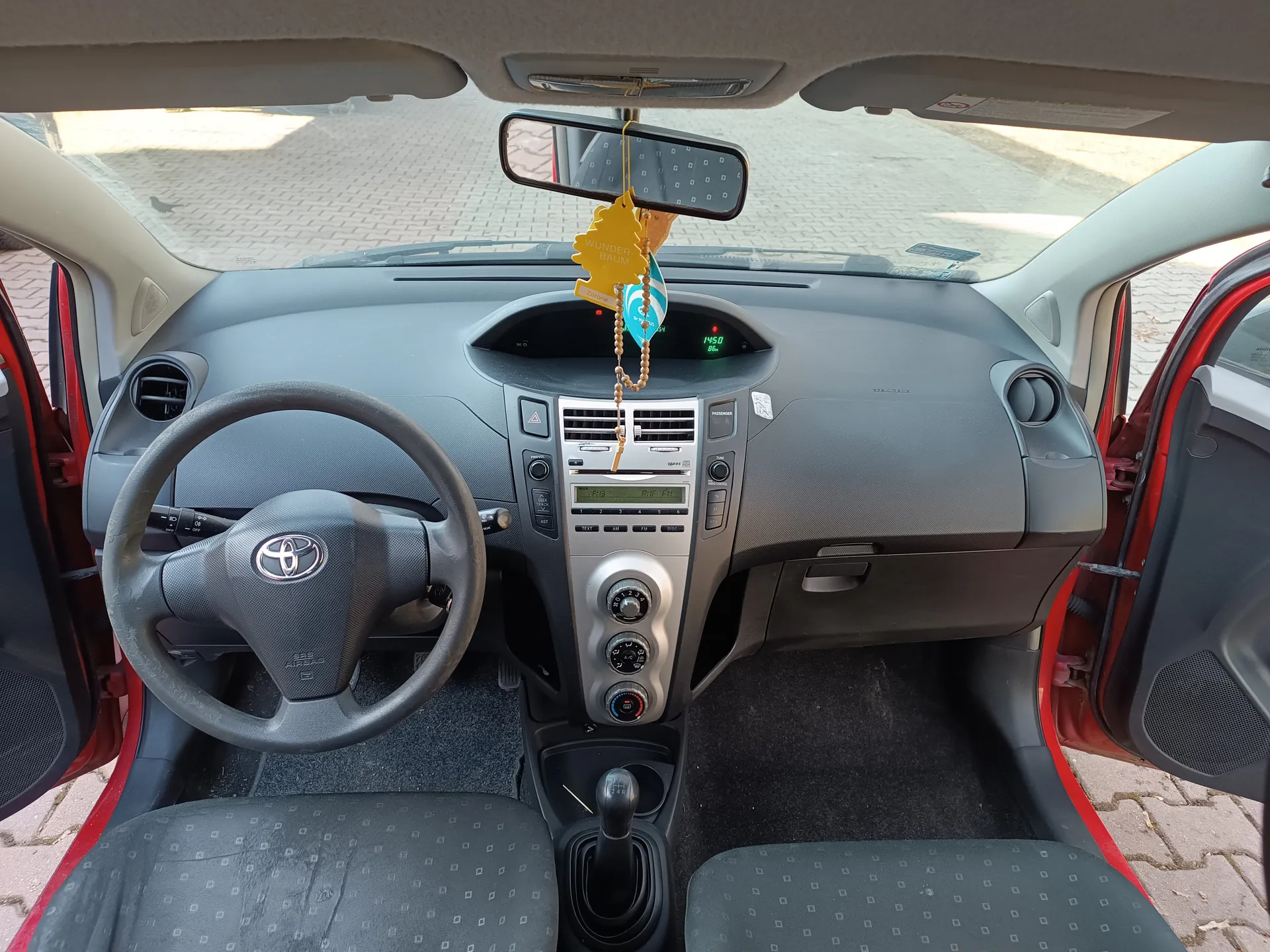 Wnętrze samochodu Toyota z widoczną kierownicą z logo marki, deska rozdzielcza z ekranem pokazującym godzinę 14:50, centralnym systemem audio i klimatyzacji. Na lusterku wstecznym wiszą żółty odświeżacz powietrza z napisem "WUNDER-BAUM" oraz brązowe koraliki. Drzwi samochodu są otwarte, odsłaniając czerwoną karoserię. Siedzenia pokryte są tkaniną z geometrycznym wzorem. Przez przednią szybę widoczny jest brukowany plac.